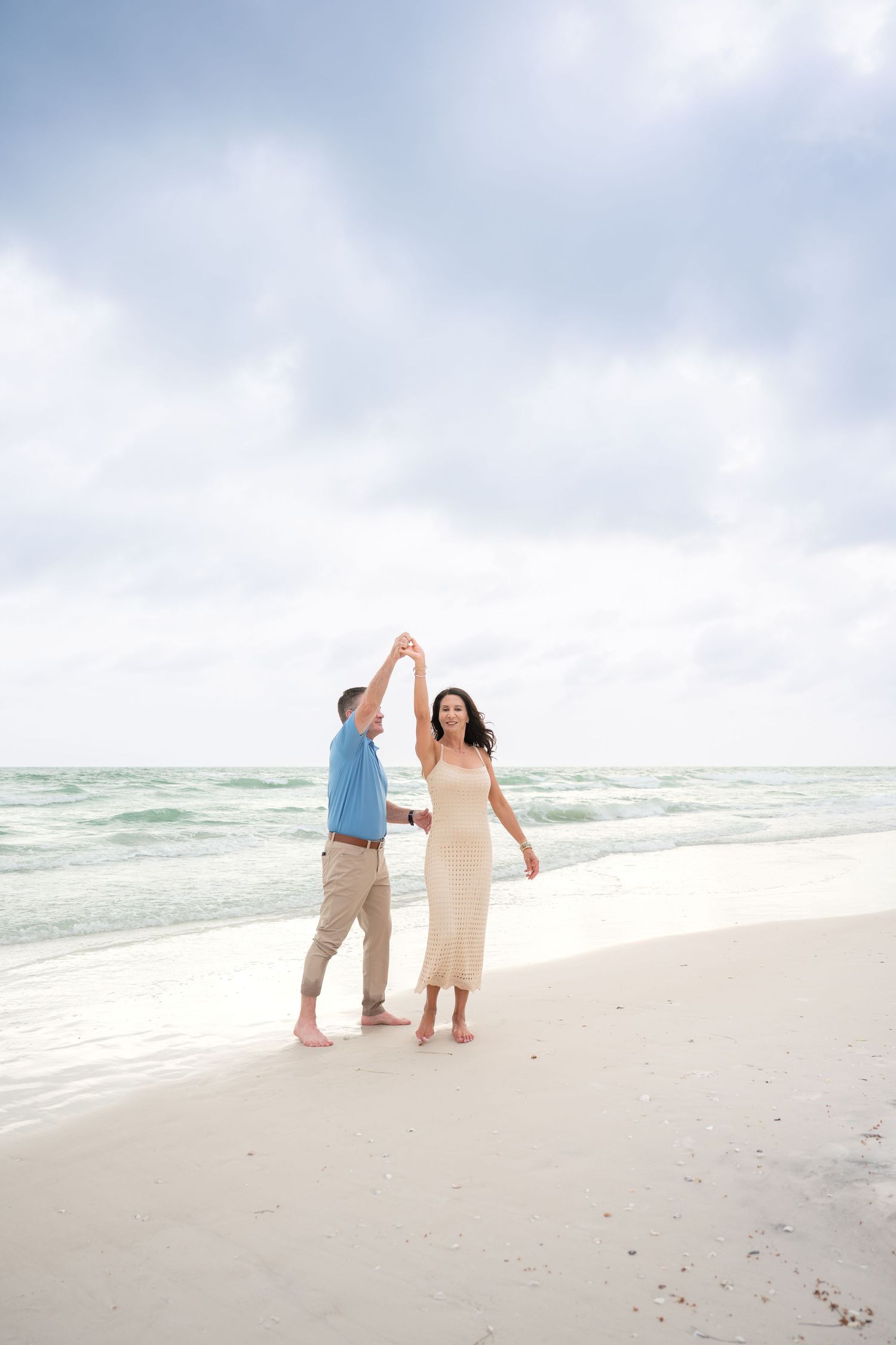 Couple dancing barefoot on a white sandy beach with turquoise ocean waves and cloudy sky backdrop.