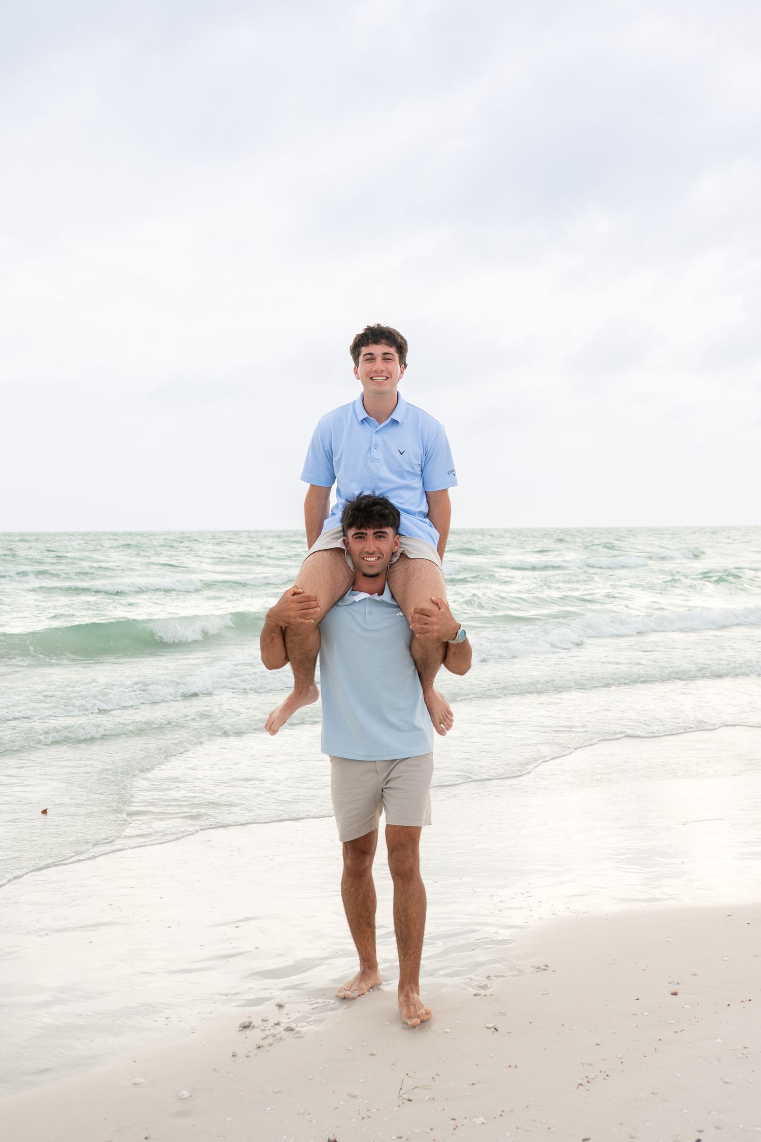 Two young men enjoy a piggyback ride on a sandy beach with turquoise ocean waves in the background.