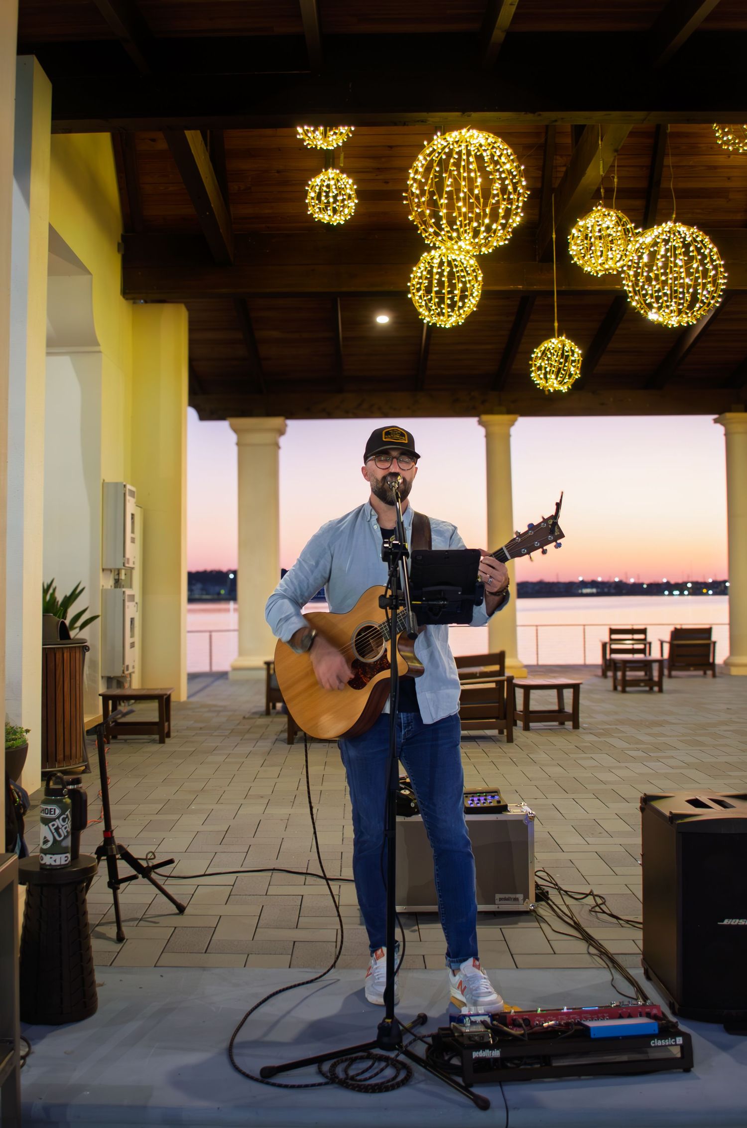 Musician performs with acoustic guitar under illuminated spherical lights at outdoor waterfront venue during sunset.
