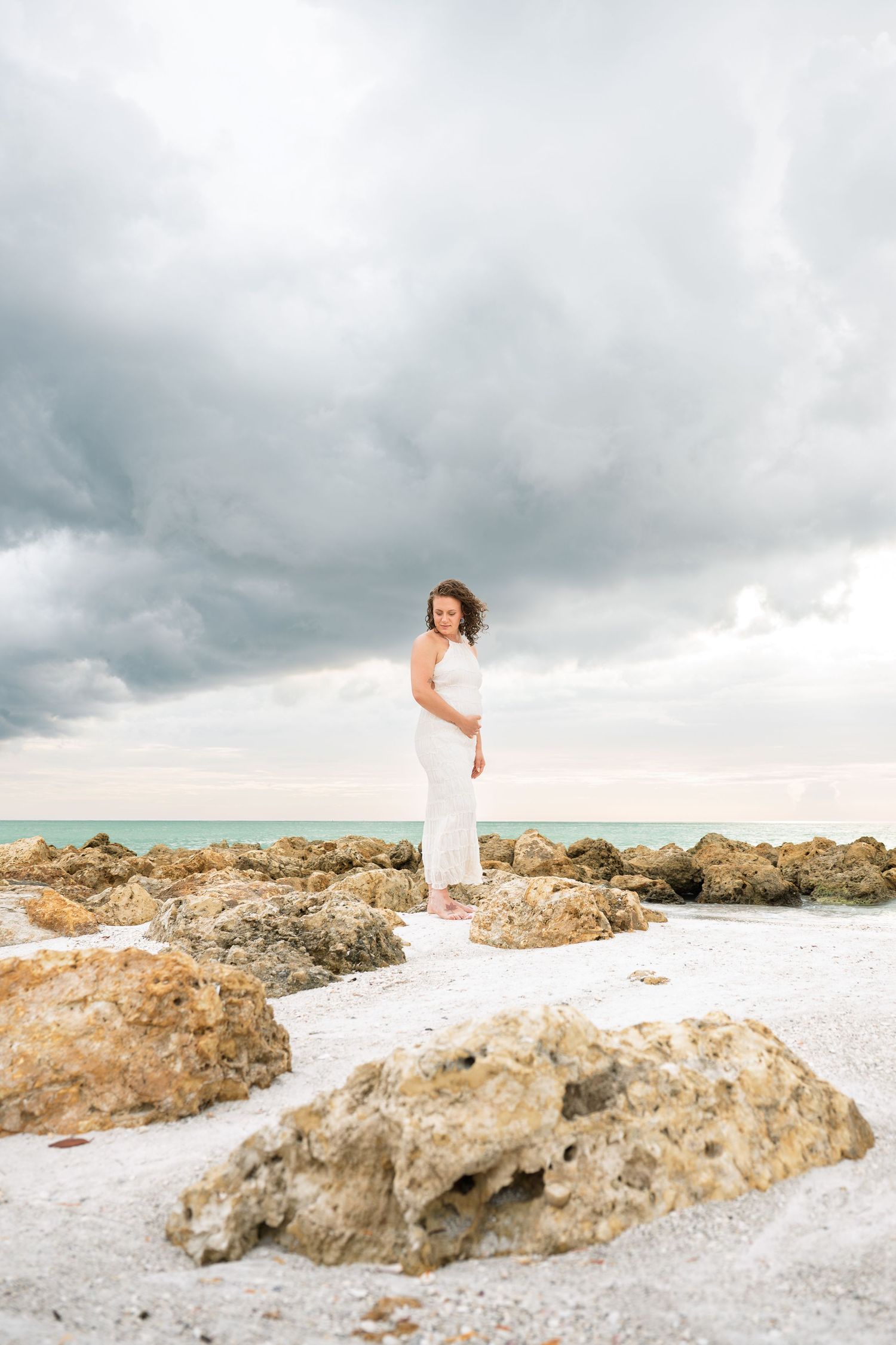Woman in white dress standing on rocky beach with dramatic cloudy sky and turquoise ocean in background.