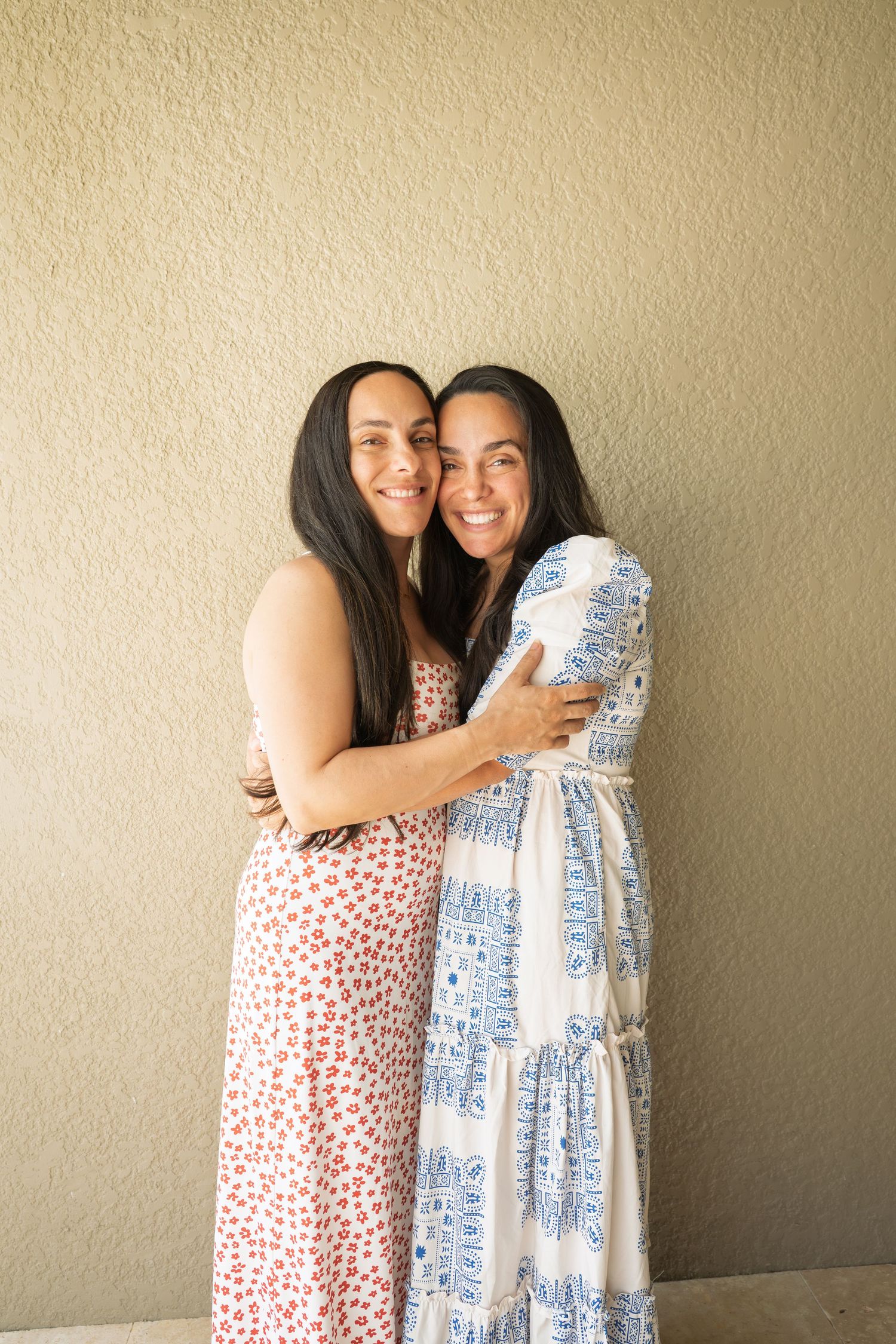 Two women in floral maxi dresses smiling and hugging against a beige wall.