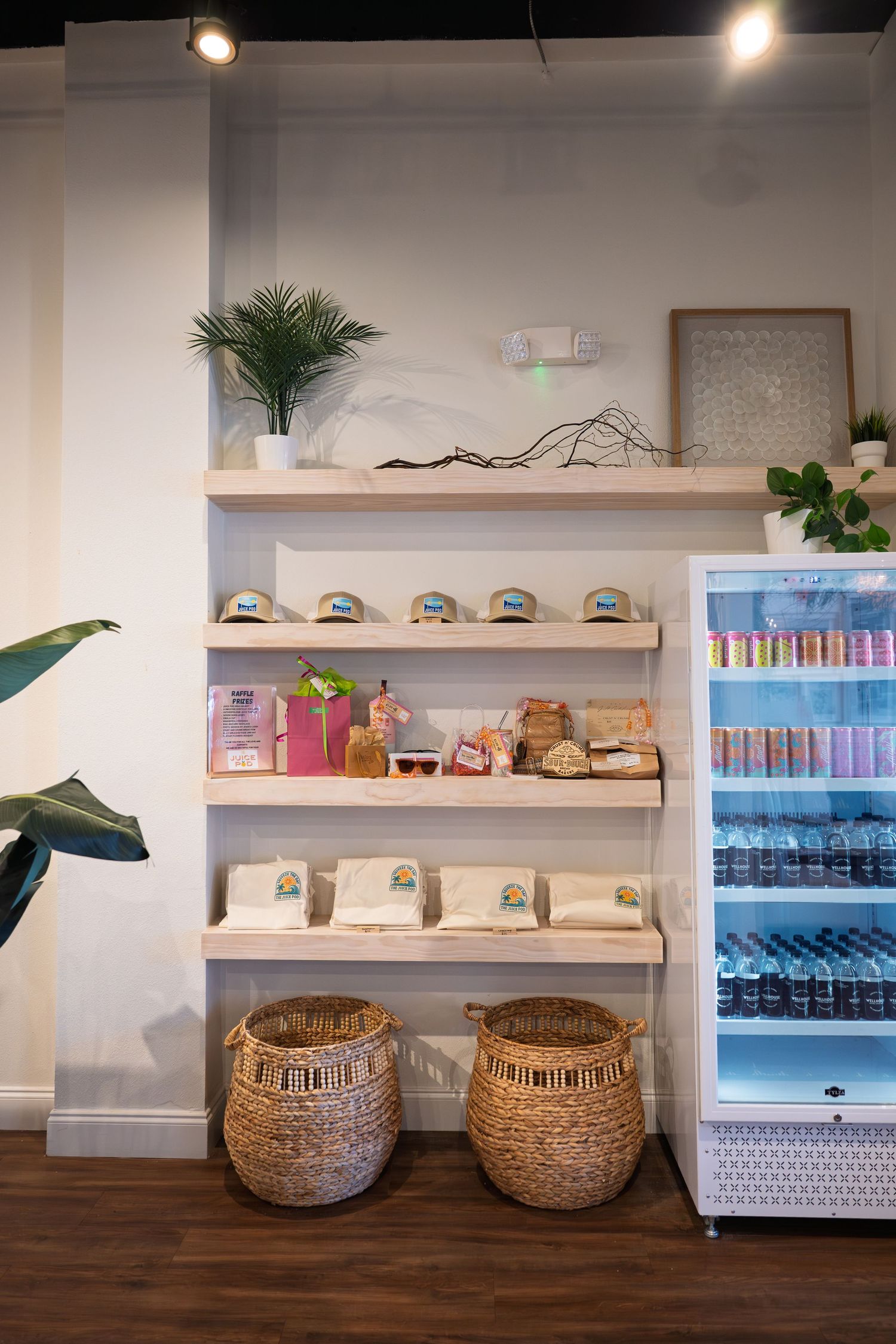 Boutique shop display with wooden shelves holding packaged goods, wicker baskets below, and a refrigerator stocked with drinks.