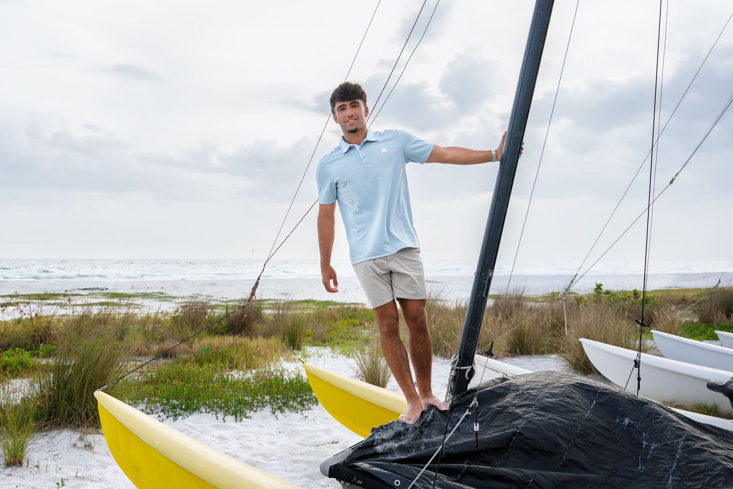 Man in light blue polo and khaki shorts stands on a sailboat hull on a sandy beach with ocean in background.