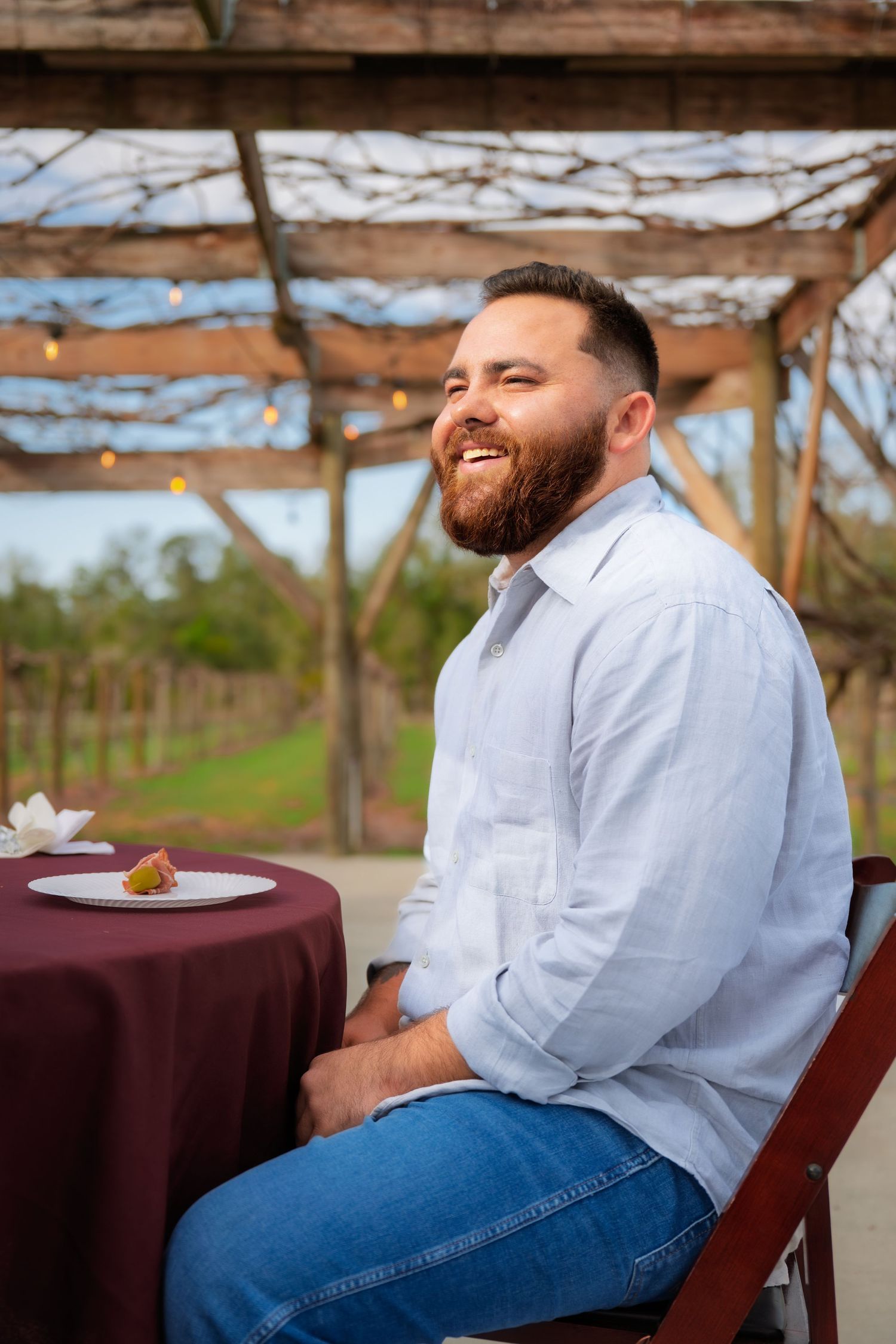 Person in light blue shirt and jeans sitting at table outdoors with burgundy tablecloth.