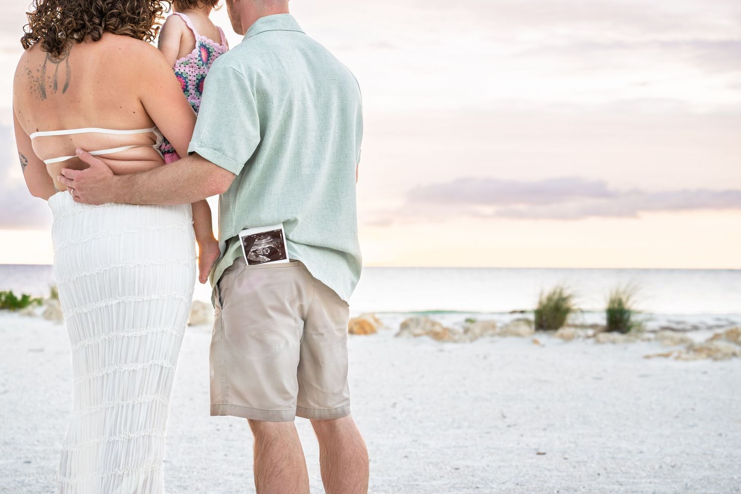 Couple on beach holding ultrasound photo, announcing pregnancy at sunset with child, romantic maternity announcement.