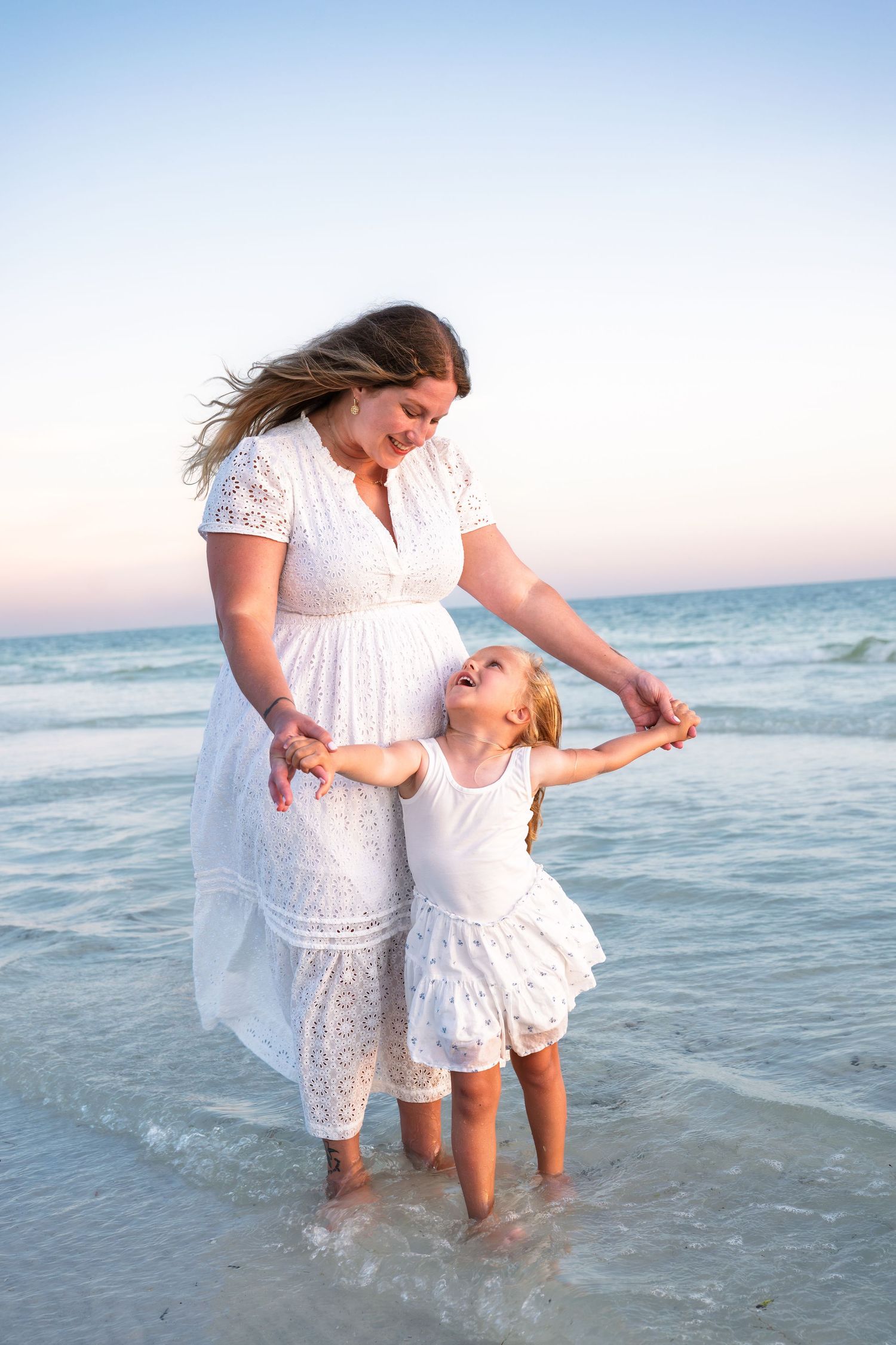 Pregnant mother and daughter in matching white dresses dancing on a beach at sunset.