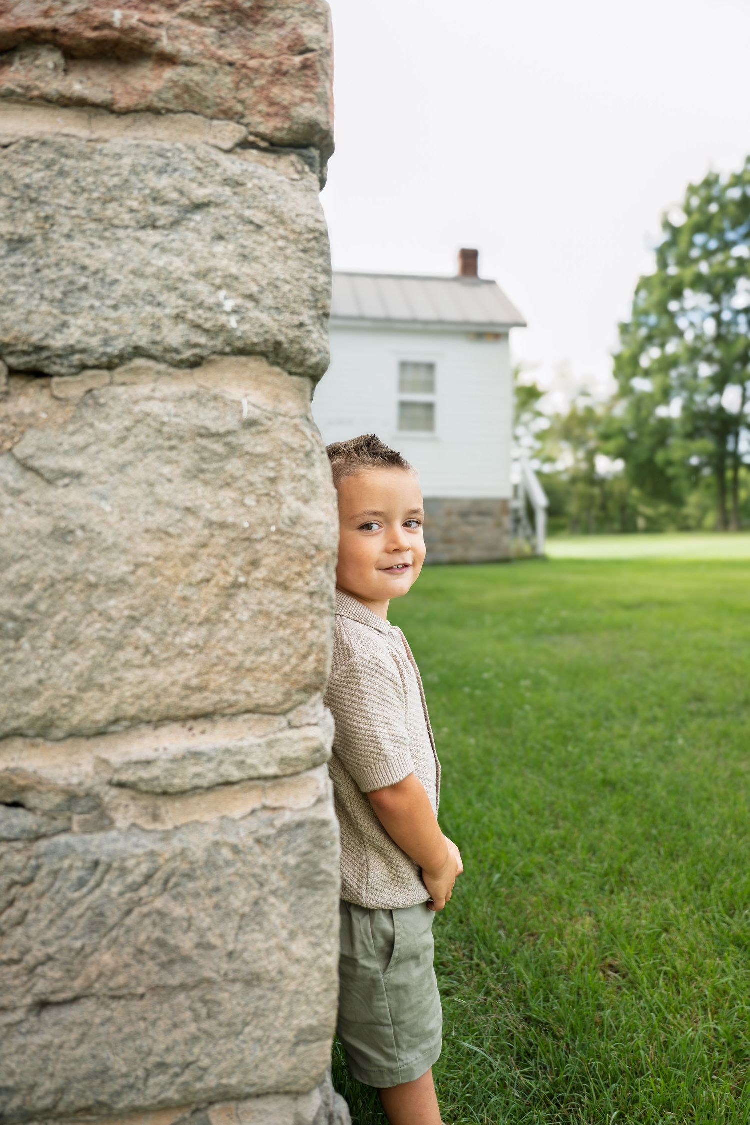 Young boy peeking around a stone pillar in a green yard with a white farmhouse in the background.