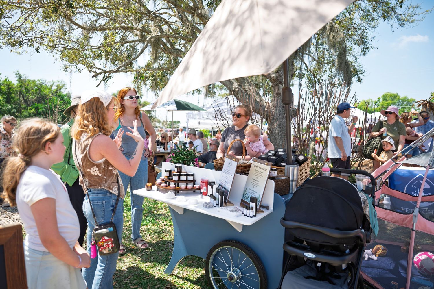 Outdoor farmers market vendor cart with artisan products displayed, surrounded by shoppers under shady trees on a sunny day.