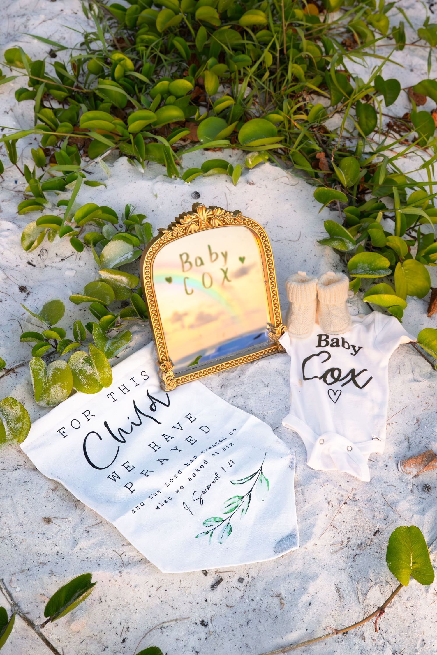 Baby Cox pregnancy announcement with onesie, mirror, and prayer sign on white sand beach surrounded by greenery.