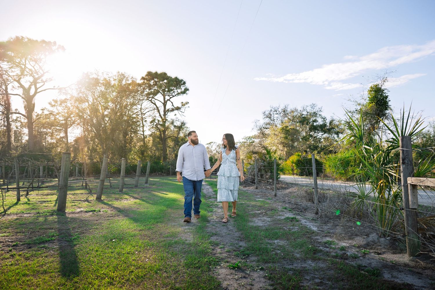 A couple walks together through a sunlit garden path surrounded by lush greenery and trees at golden hour.