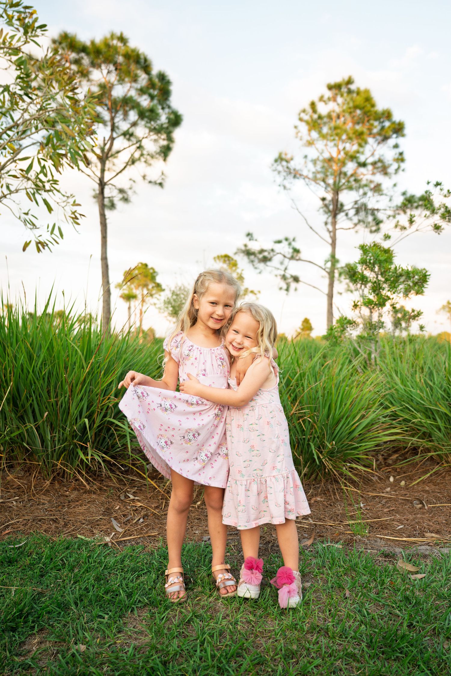 Two young blonde girls in matching pink floral dresses hugging outdoors with green trees and grass in background.