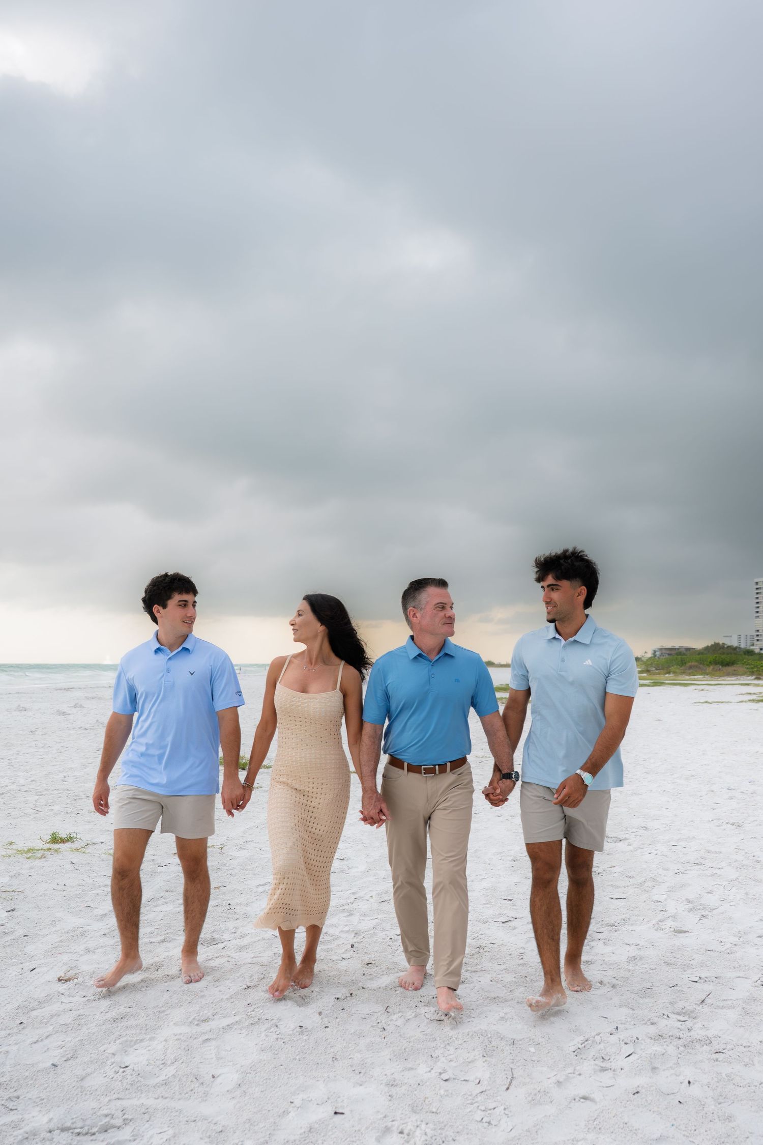 Family of four walks hand-in-hand on white sand beach under cloudy skies, wearing coordinated blue and beige outfits.