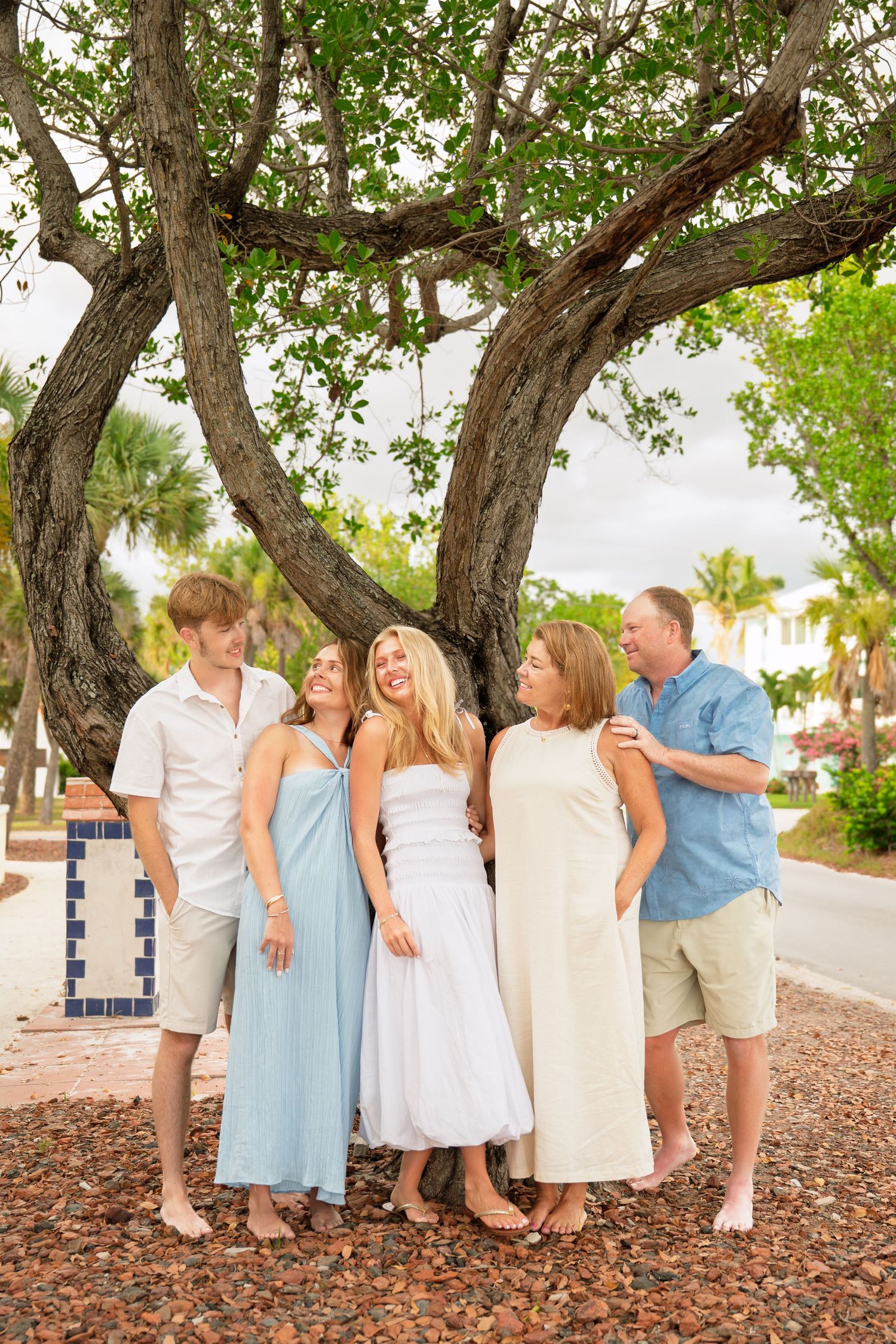 Family of five posing together under a large tree in a tropical setting, wearing coordinated light-colored outfits.