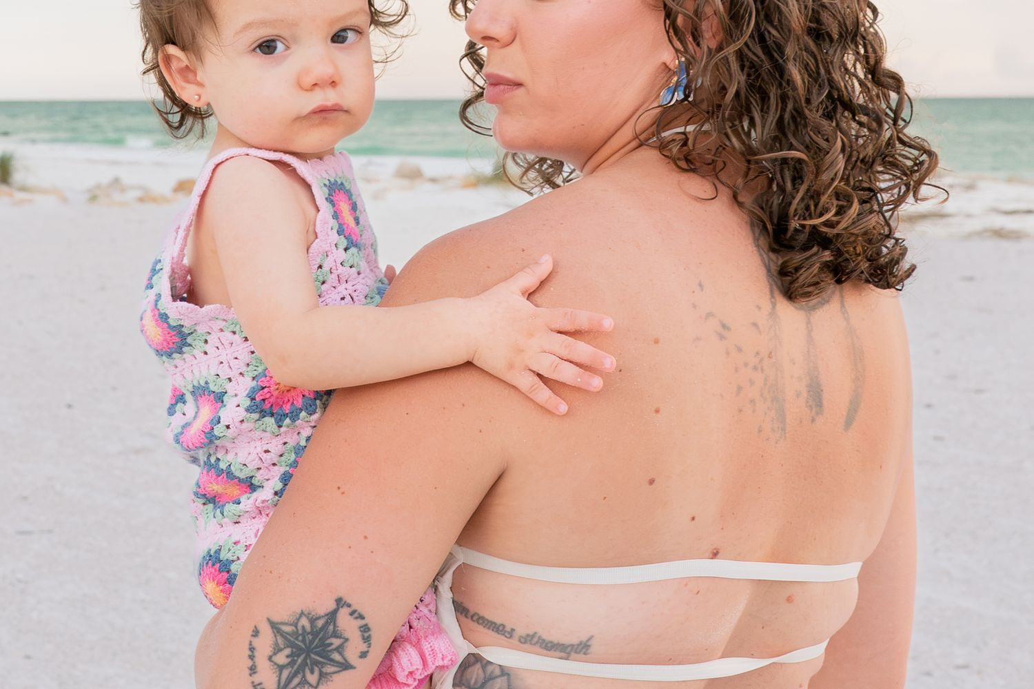 Mother with curly hair and tattoos holds baby girl in colorful swimsuit on a sandy beach.