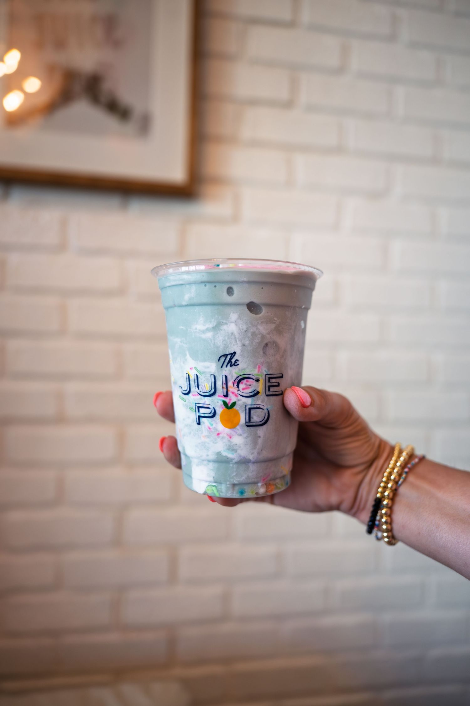 A hand holding a colorful The Juice Pod drink cup against a white brick wall background.