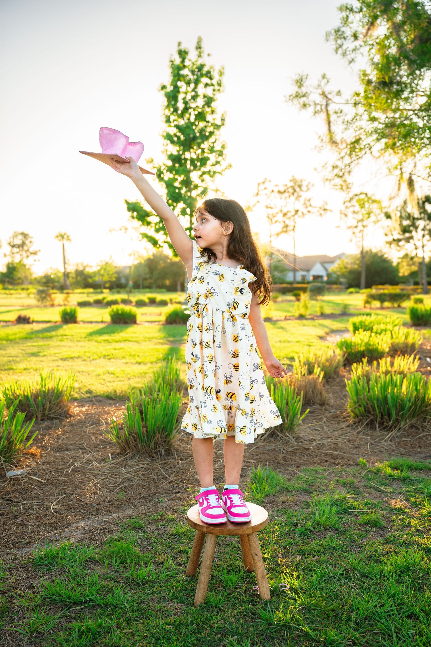 Young girl in floral dress stands on wooden stool, holding pink paper airplane aloft in sunny green garden.