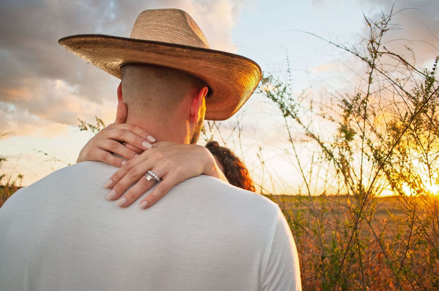 A couple embraces in a golden wheat field during sunset with sunflare and soft lighting.