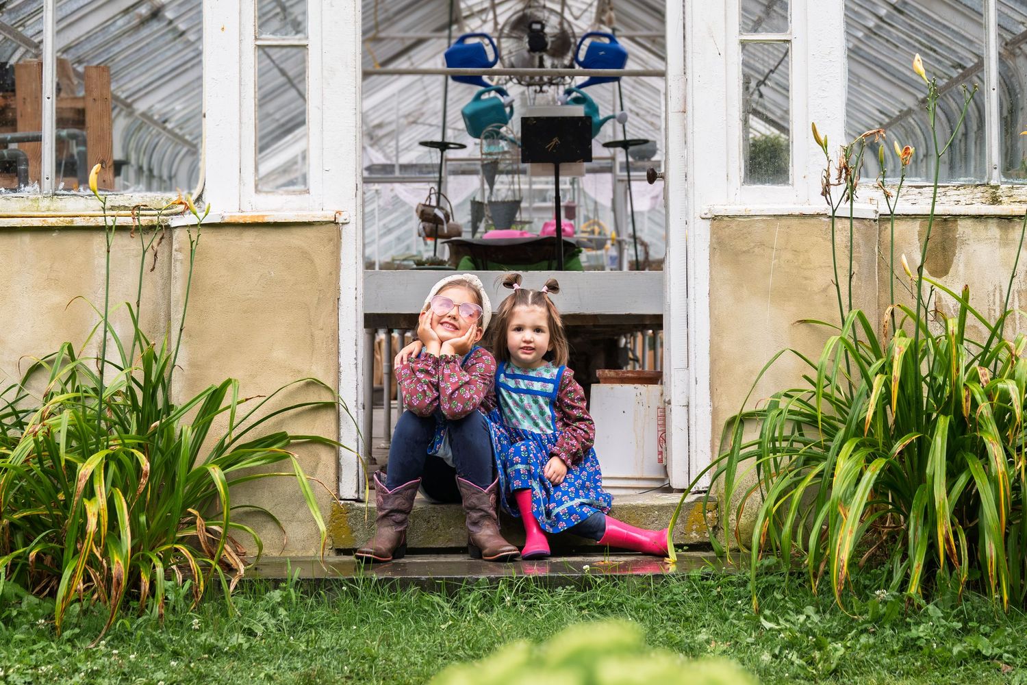 Two children sitting and smiling in the doorway of a white greenhouse surrounded by green plants.