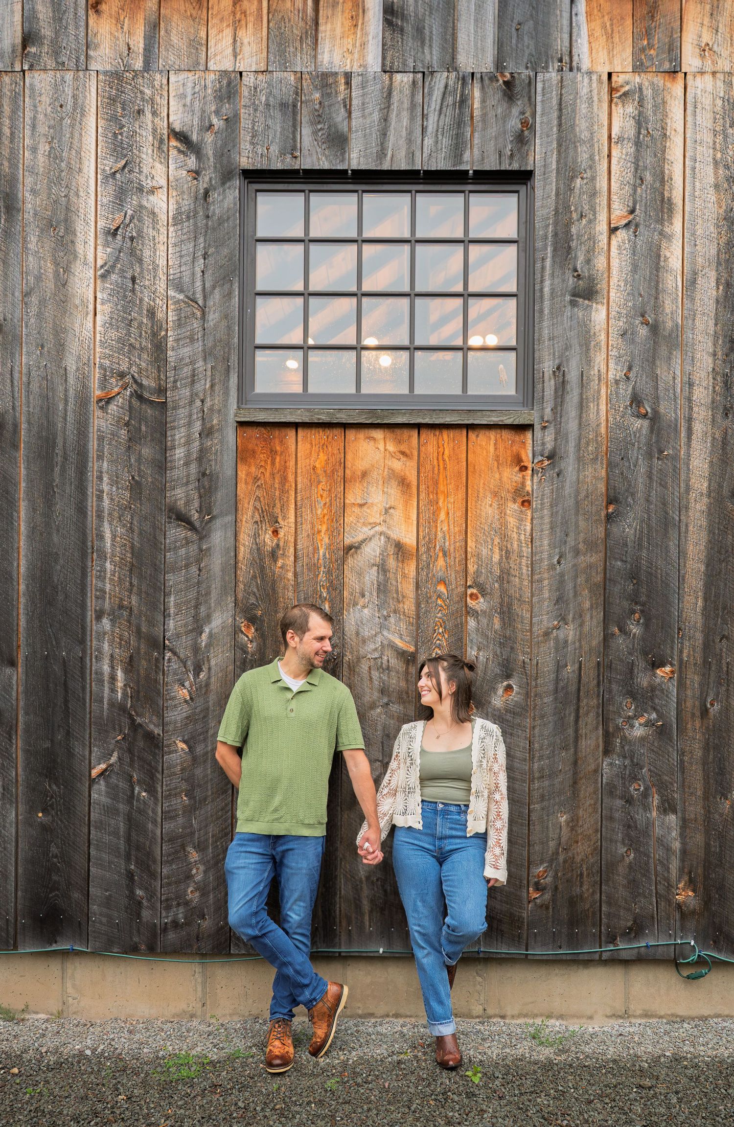 Couple holding hands and smiling at each other against a rustic wooden barn wall with a window above them.