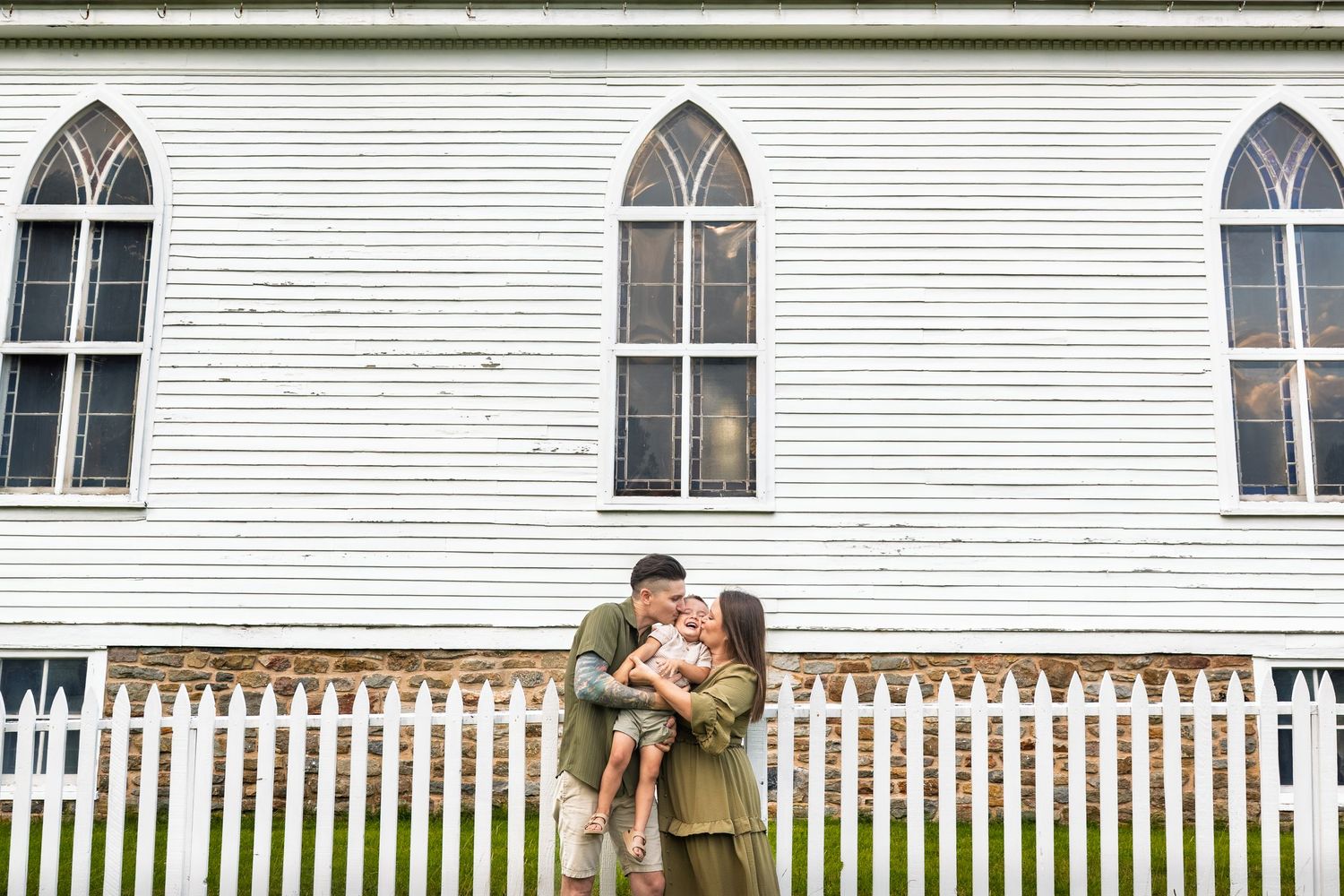 A couple shares a tender moment with their baby in front of a white church with gothic windows and picket fence.