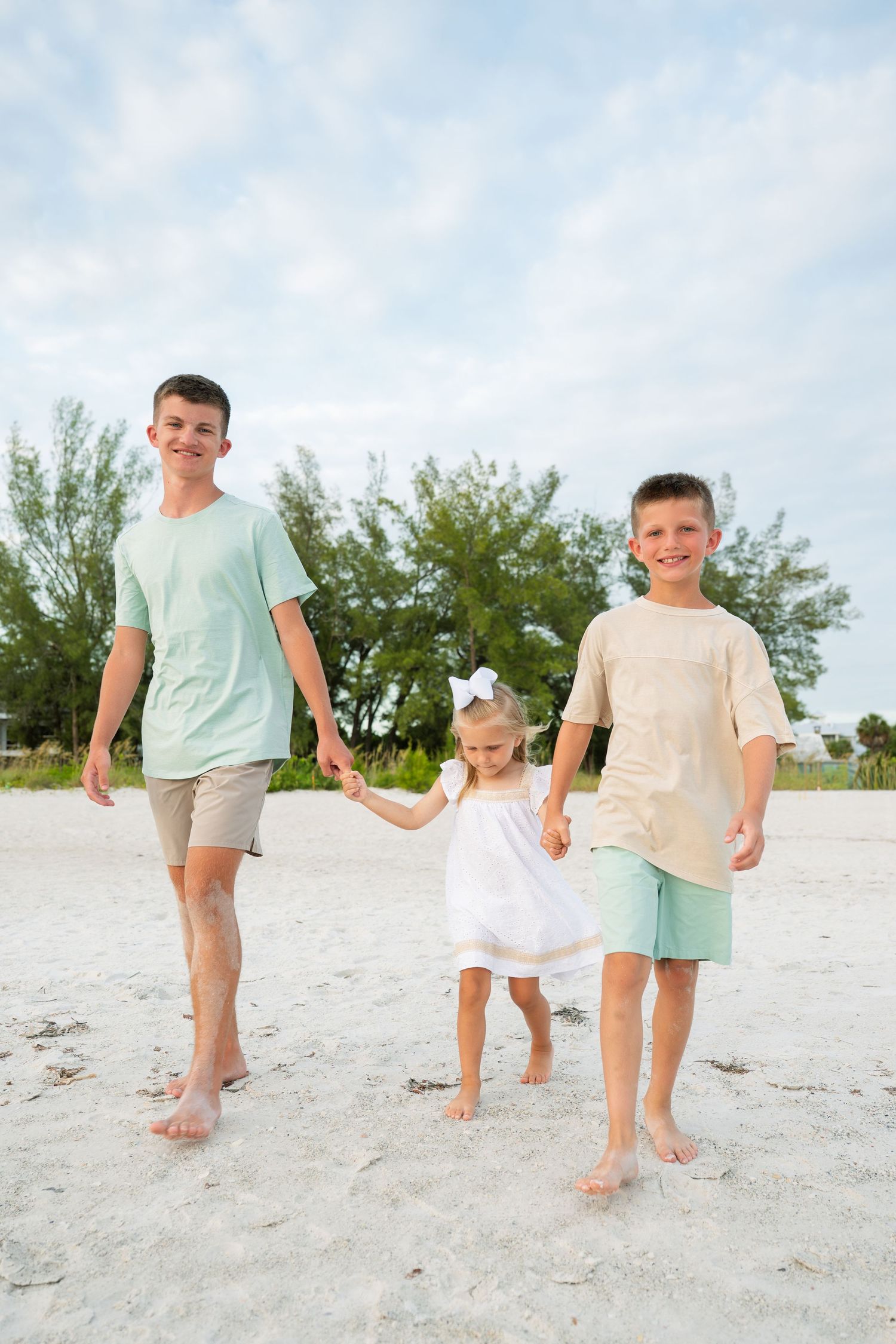 Two boys and a toddler girl in white dress hold hands walking barefoot on a sandy beach with trees in background.