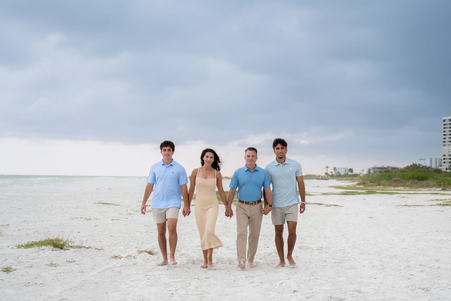 Family of four walks hand-in-hand on a white sandy beach under cloudy skies near a coastal resort.
