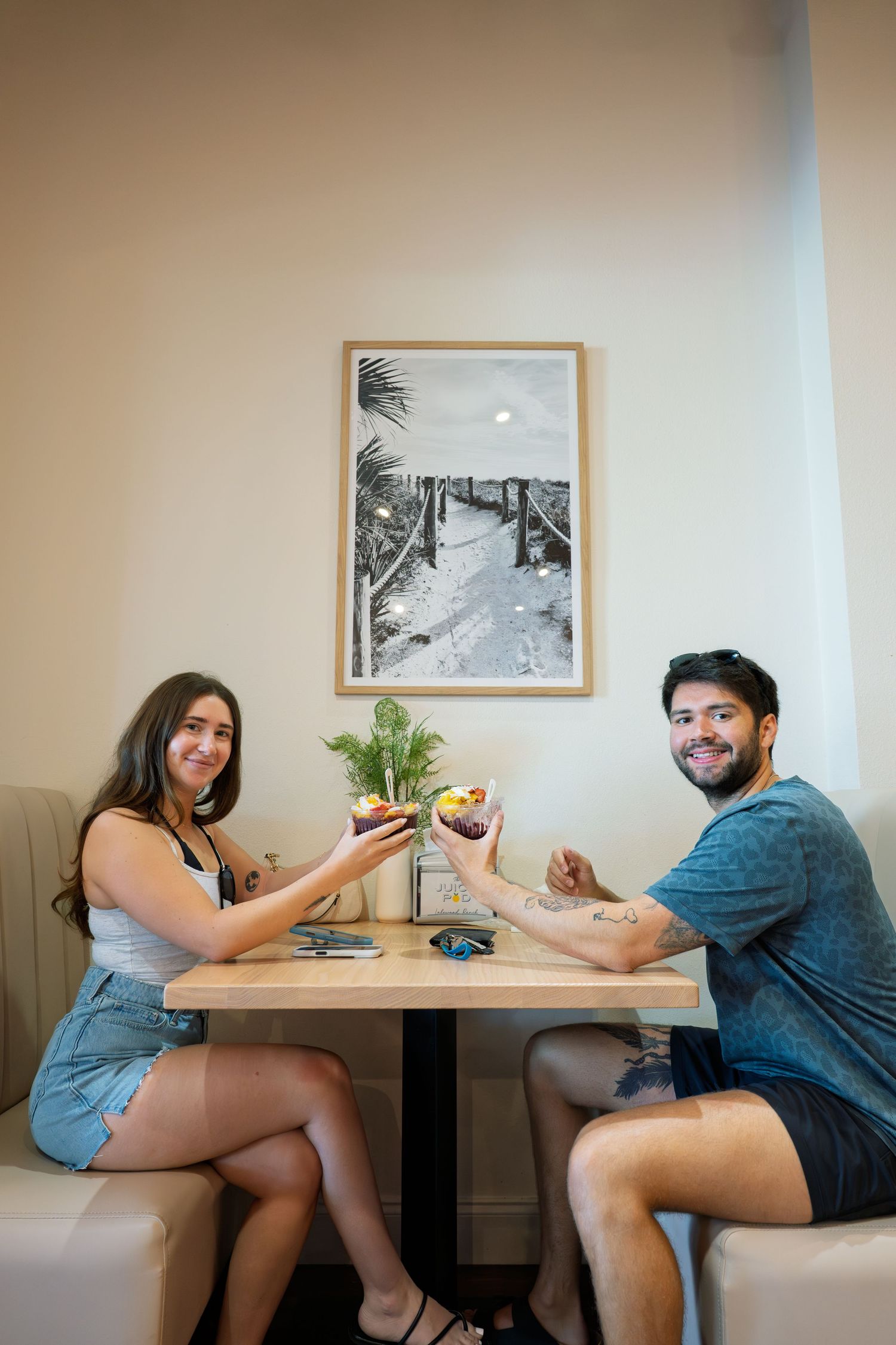 Couple smiling and toasting with colorful bowls at a cafe table, with a black and white beach photo on the wall.