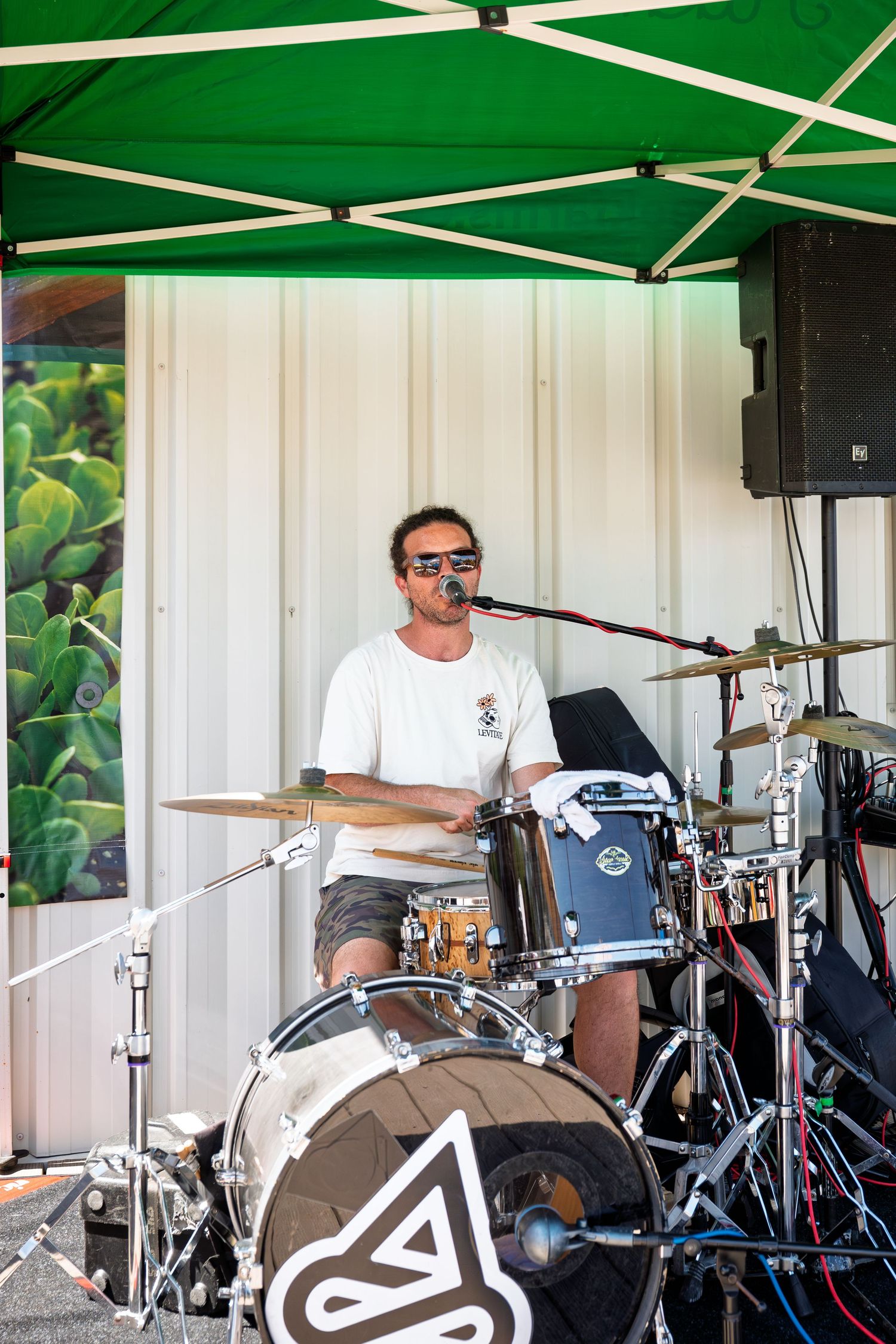 Male drummer performing on stage outdoors under a green canopy tent at a live music event.