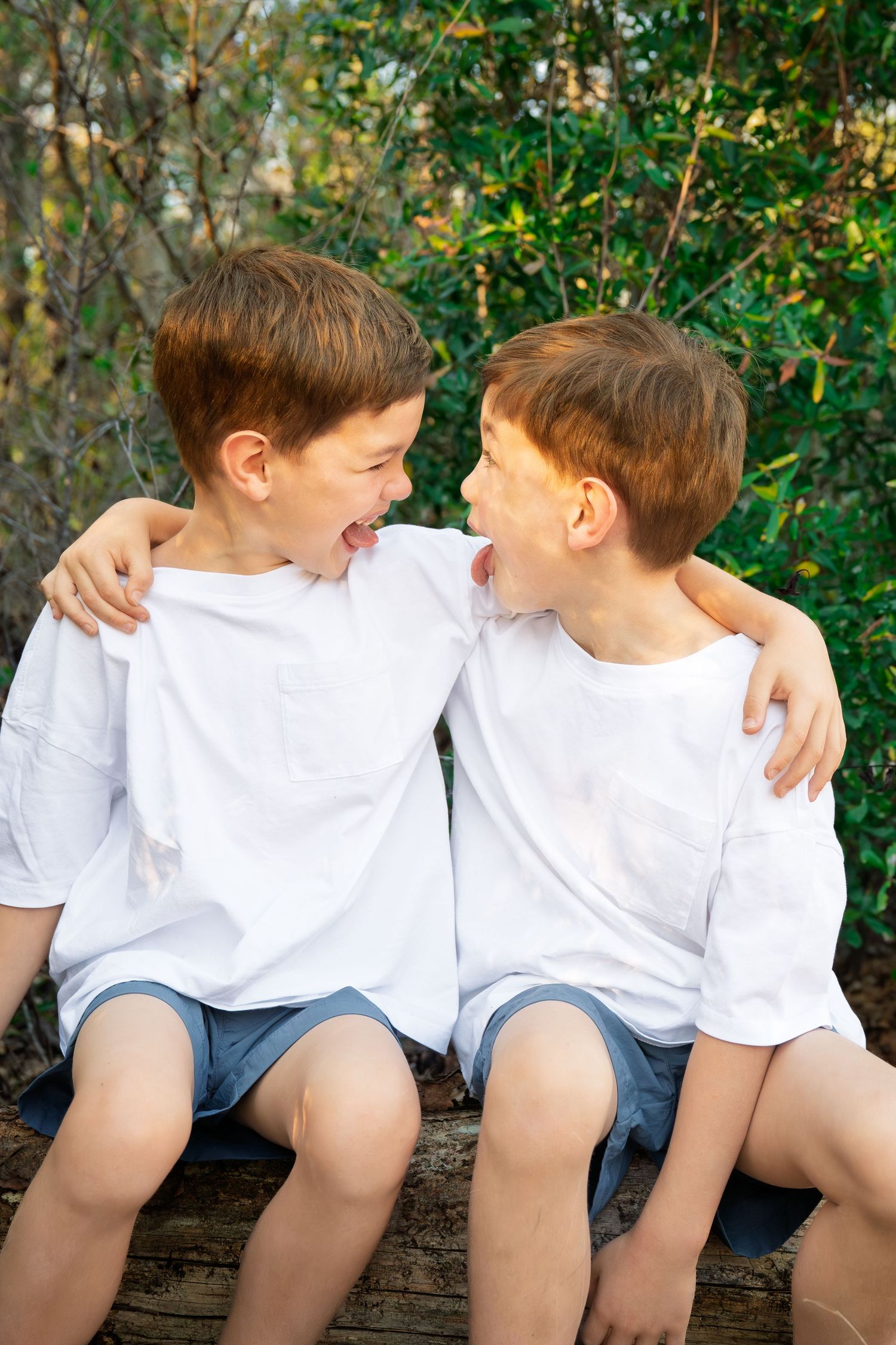 Two laughing twin boys in white shirts and blue shorts sit together outdoors, arms around each other.