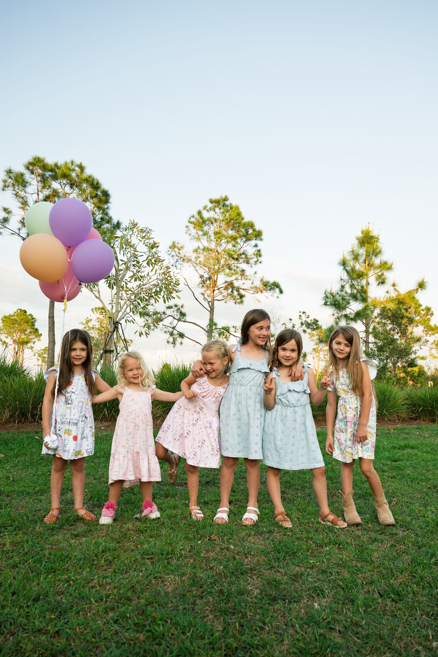 Six young girls in colorful dresses holding balloons together outdoors.