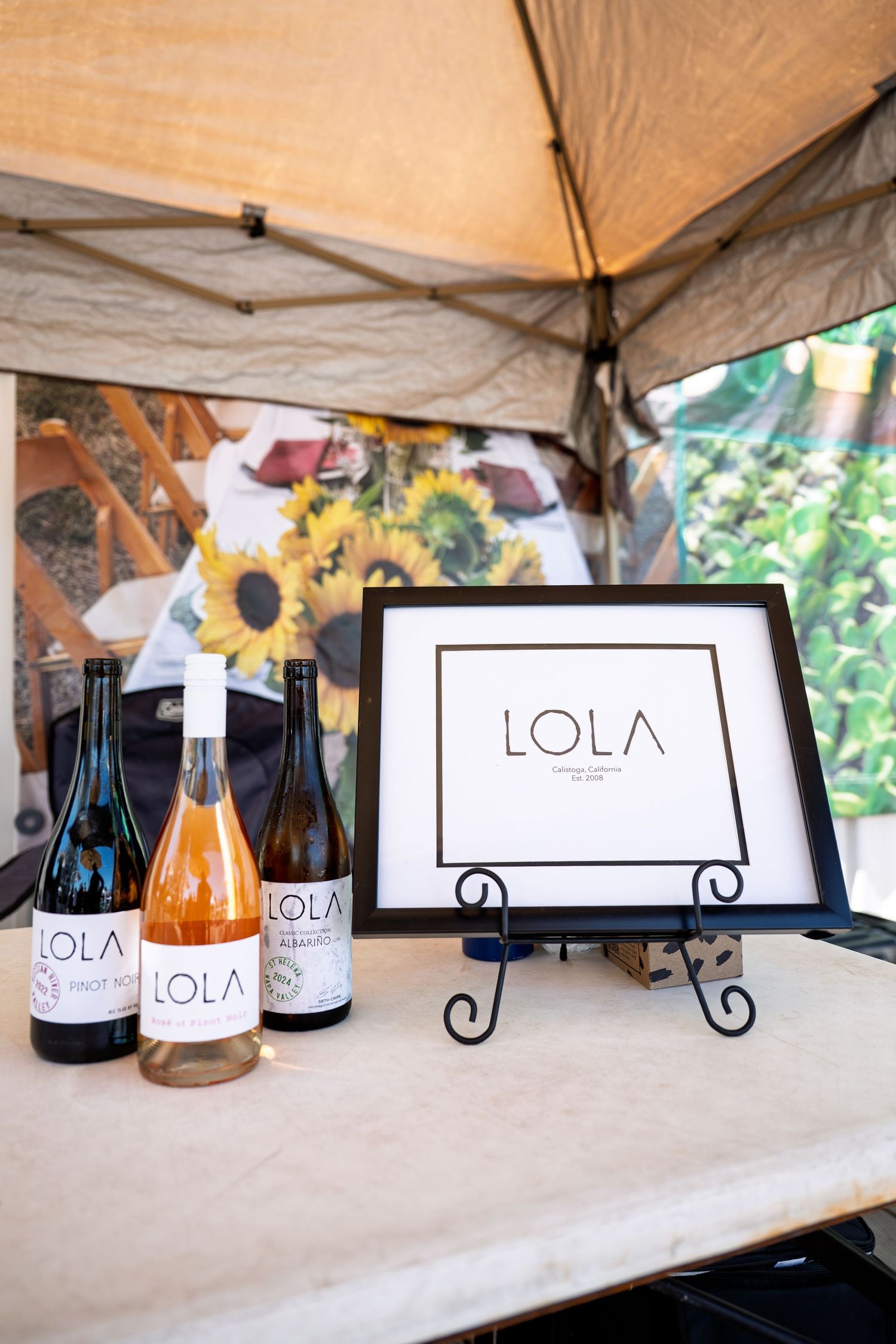 Three LOLA wine bottles displayed beside a framed LOLA logo sign under a tent with sunflowers in the background.