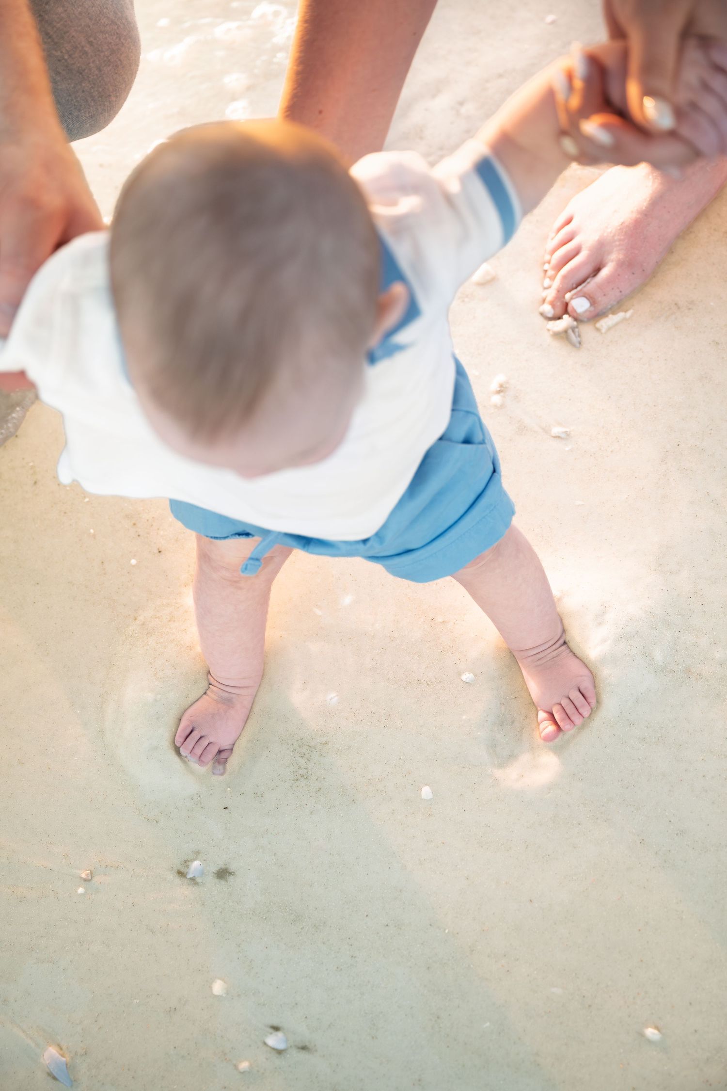 Baby standing on sandy beach with bare feet, wearing blue shorts and white bib, held by adult hands.