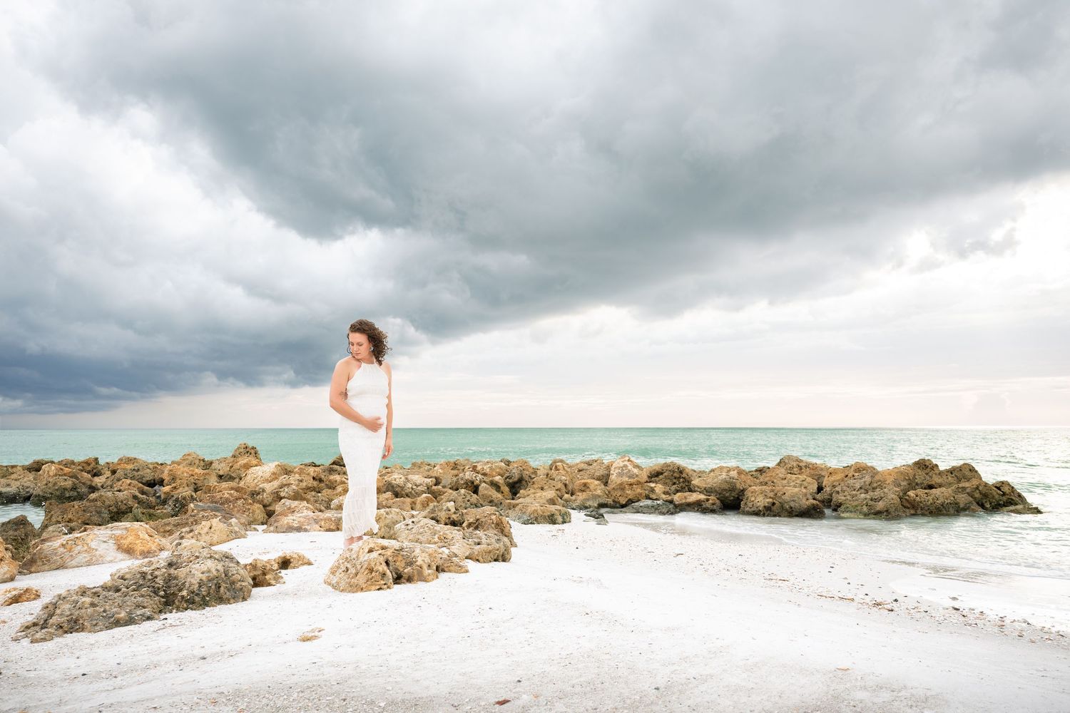 Bride in white gown stands on rocky beach under dramatic stormy sky with turquoise ocean waters in background.