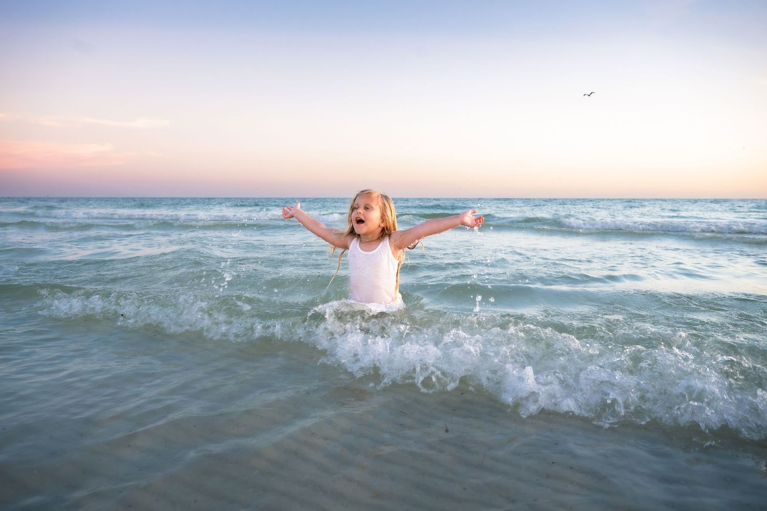 Young girl in white dress joyfully playing in ocean waves at sunset with arms outstretched.