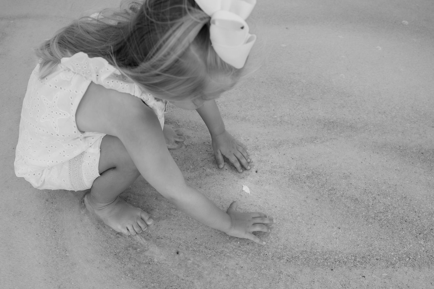 Black and white photo of a young girl in a white dress and hat playing in the sand at the beach.