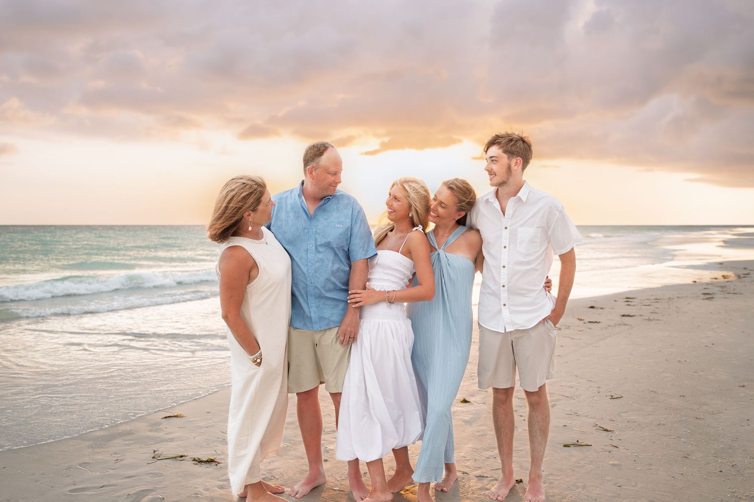 Family of five posing together on a sandy beach at sunset with pastel skies and ocean waves in the background.