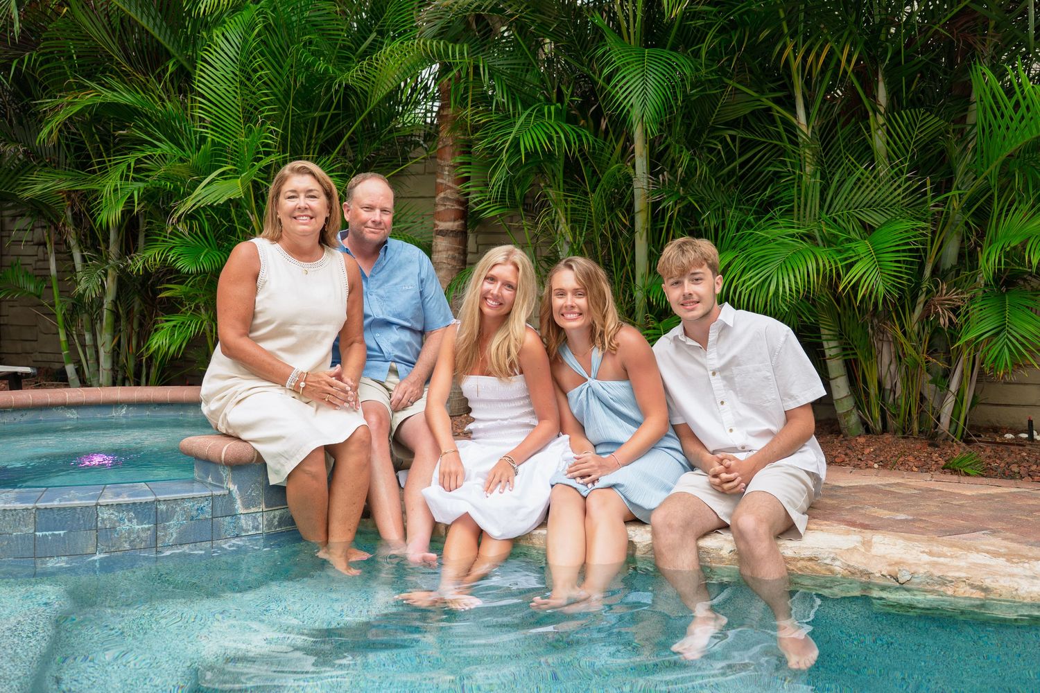 Happy family of five sitting poolside with feet in water, surrounded by tropical palm trees.