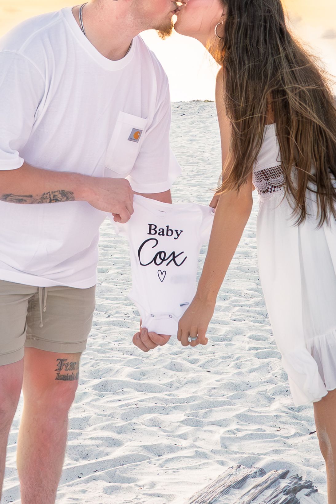 Couple kissing on beach holding Baby Cox onesie, pregnancy announcement photo on sandy shore.