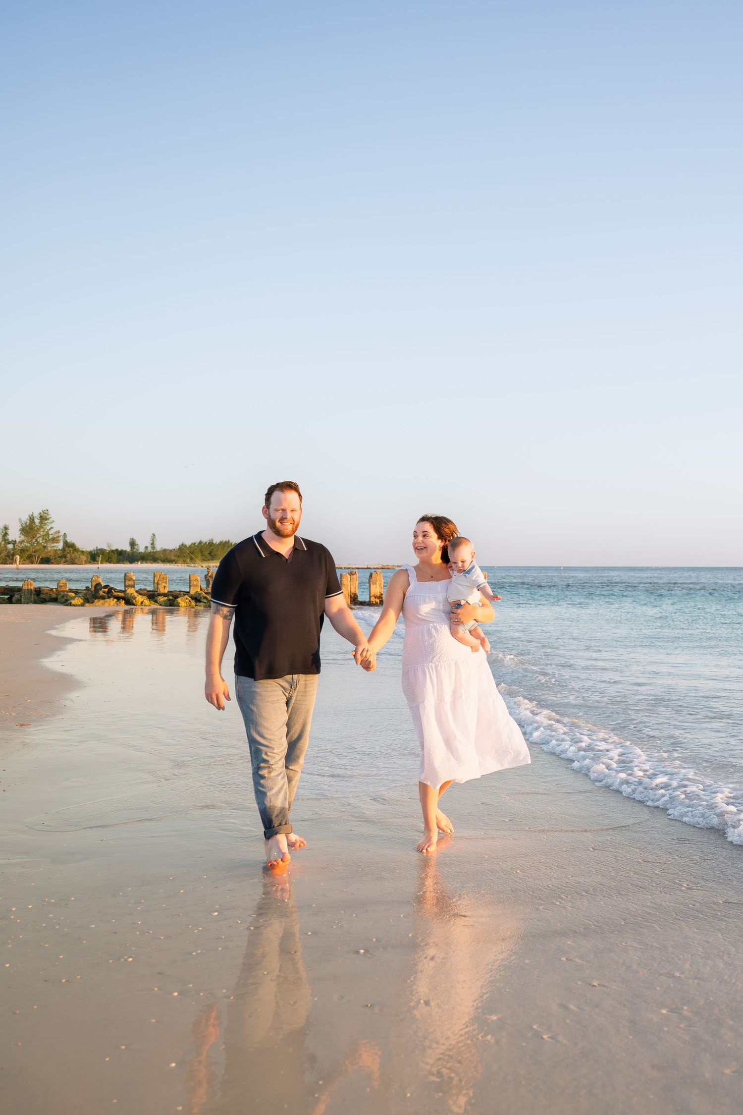 A happy family walks hand-in-hand along a sandy beach at sunset, with a woman in white holding a child.