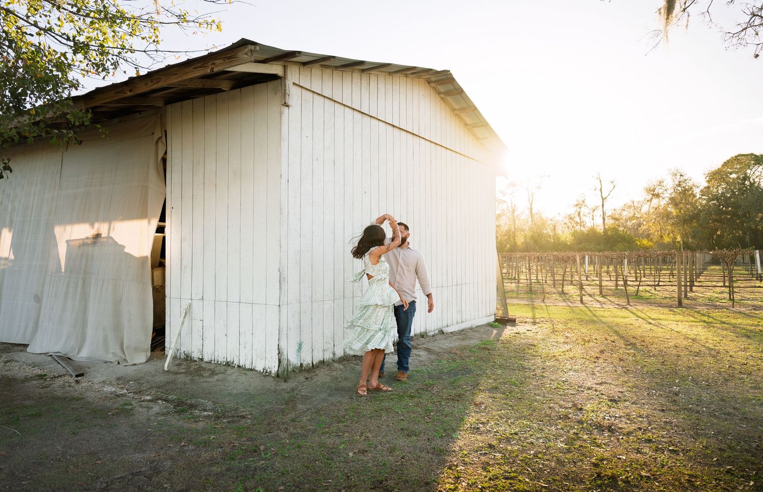A romantic sunset photo session in front of a rustic white barn with golden light streaming through the trees.