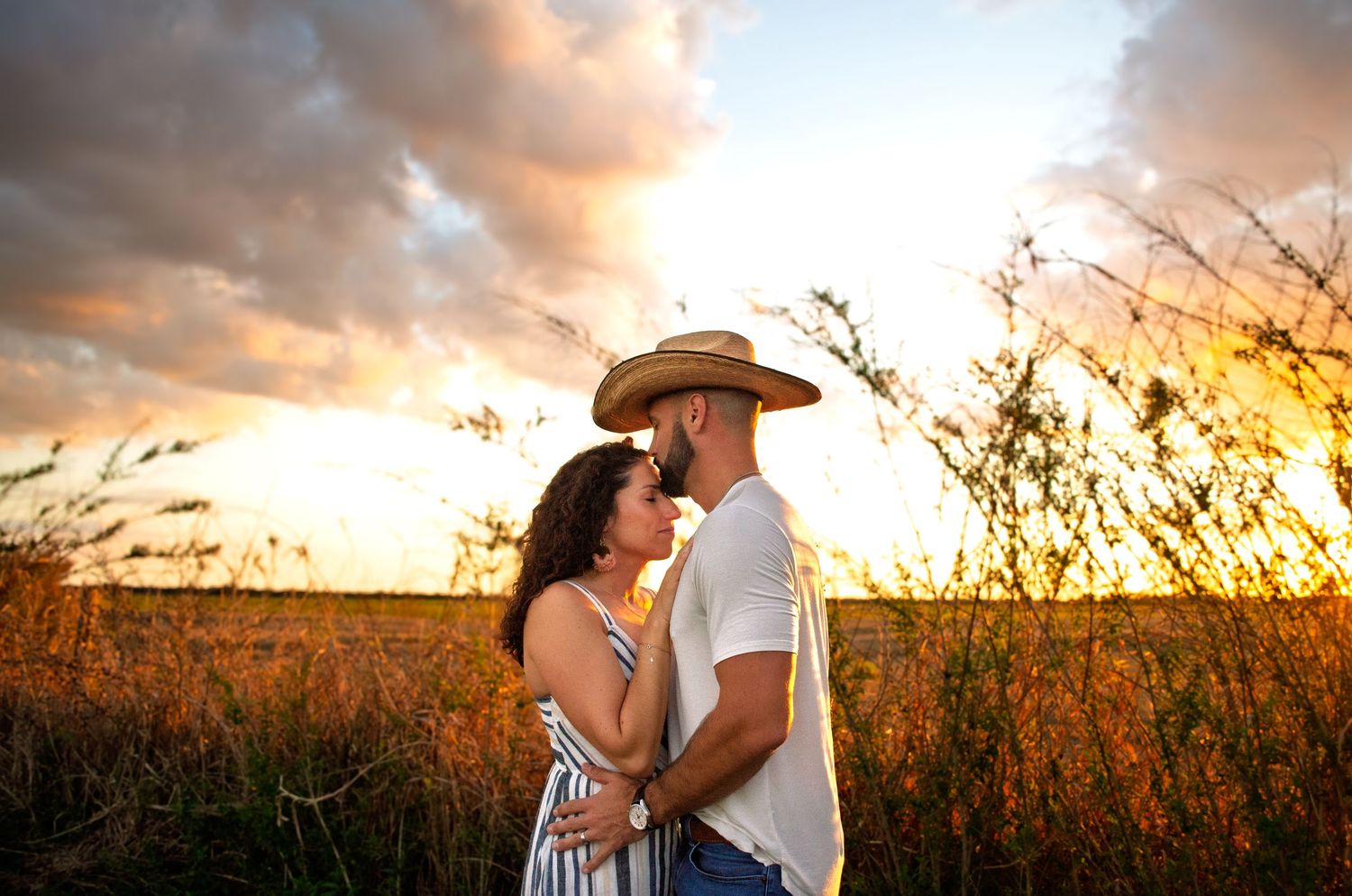 Couple embraces in golden wheat field at sunset creating romantic silhouettes across panoramic countryside view.