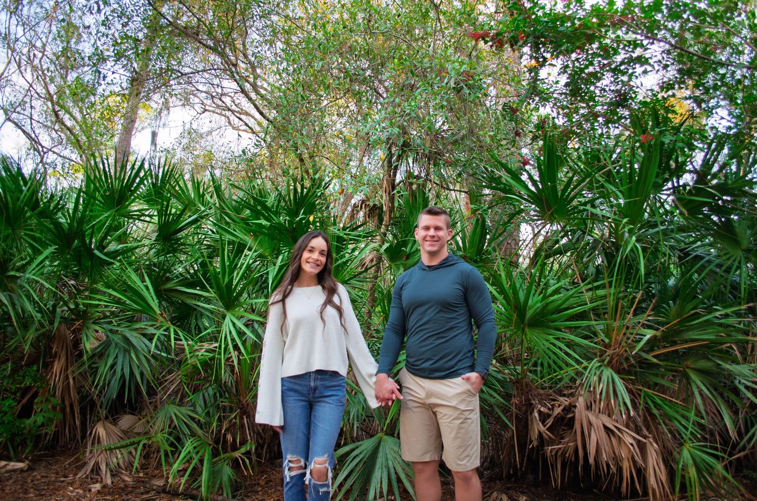 A couple poses together in front of lush tropical palm plants at a botanical garden.