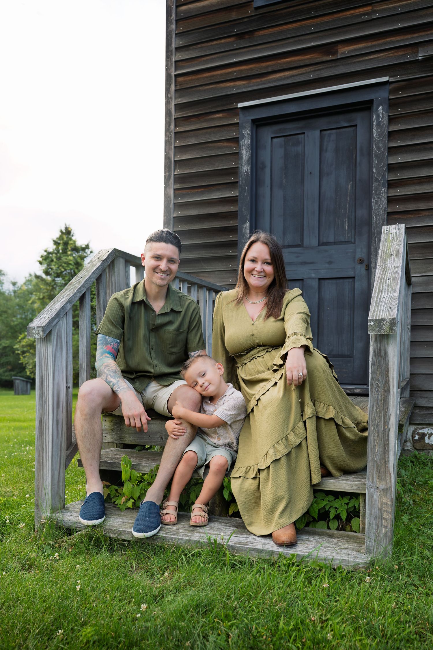 Family of three sitting on wooden steps of rustic building, parents in green outfits with toddler between them.