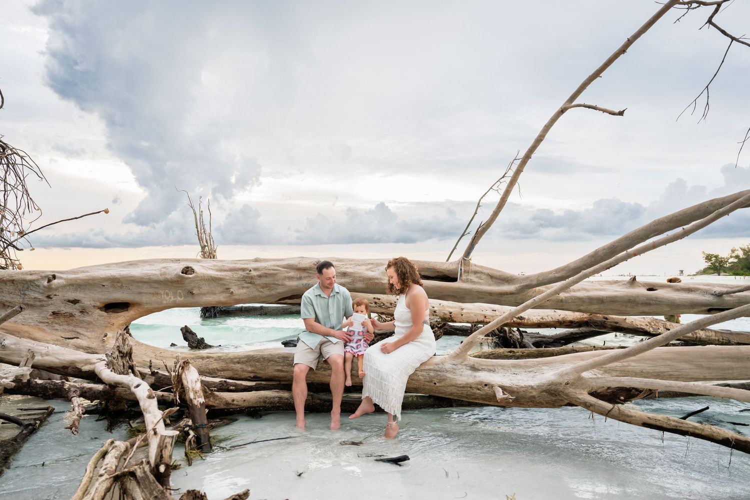 Couple sitting on driftwood logs on a beach with dramatic cloudy sky backdrop.