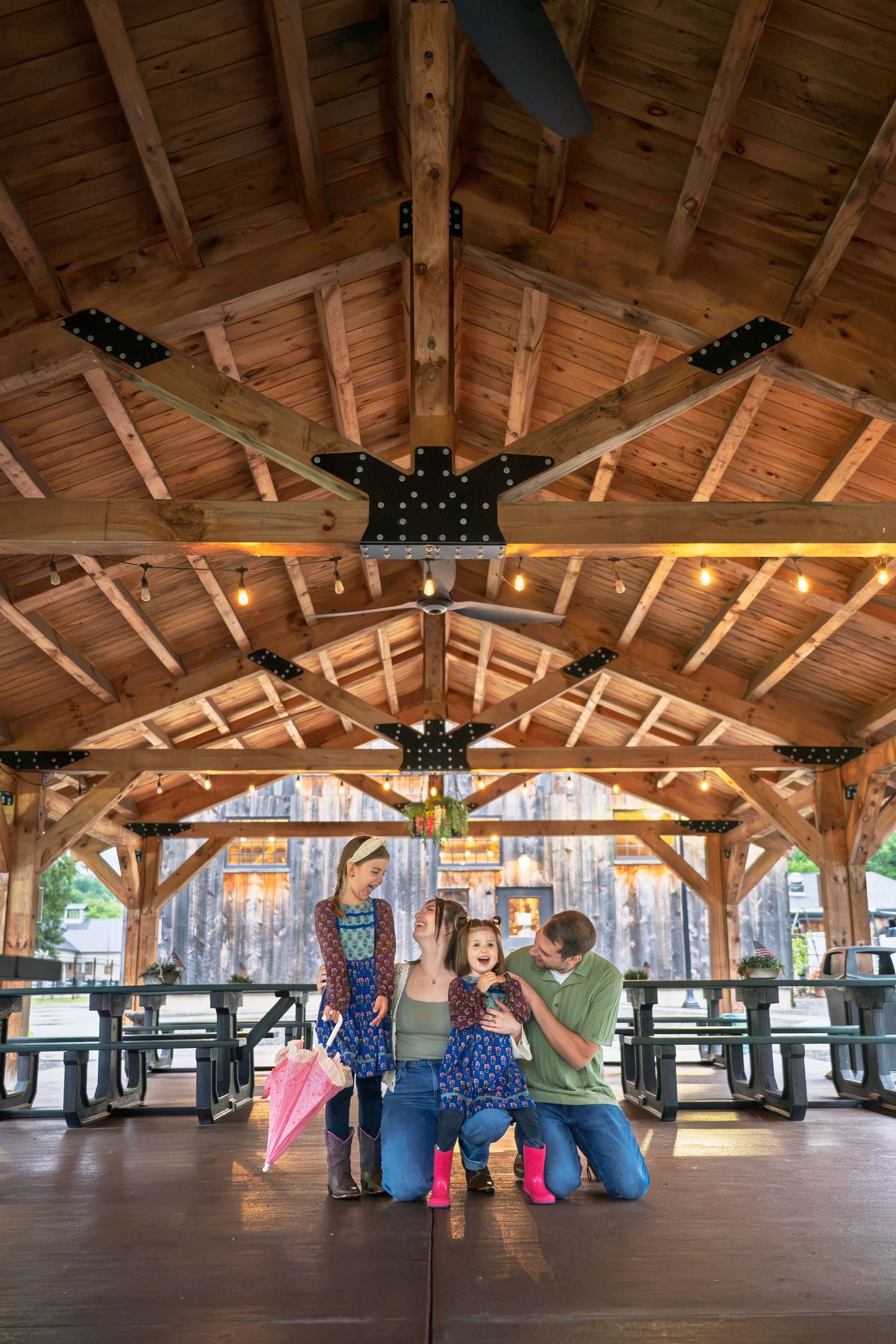Family of four enjoying time together under a rustic wooden pavilion with string lights and picnic tables.