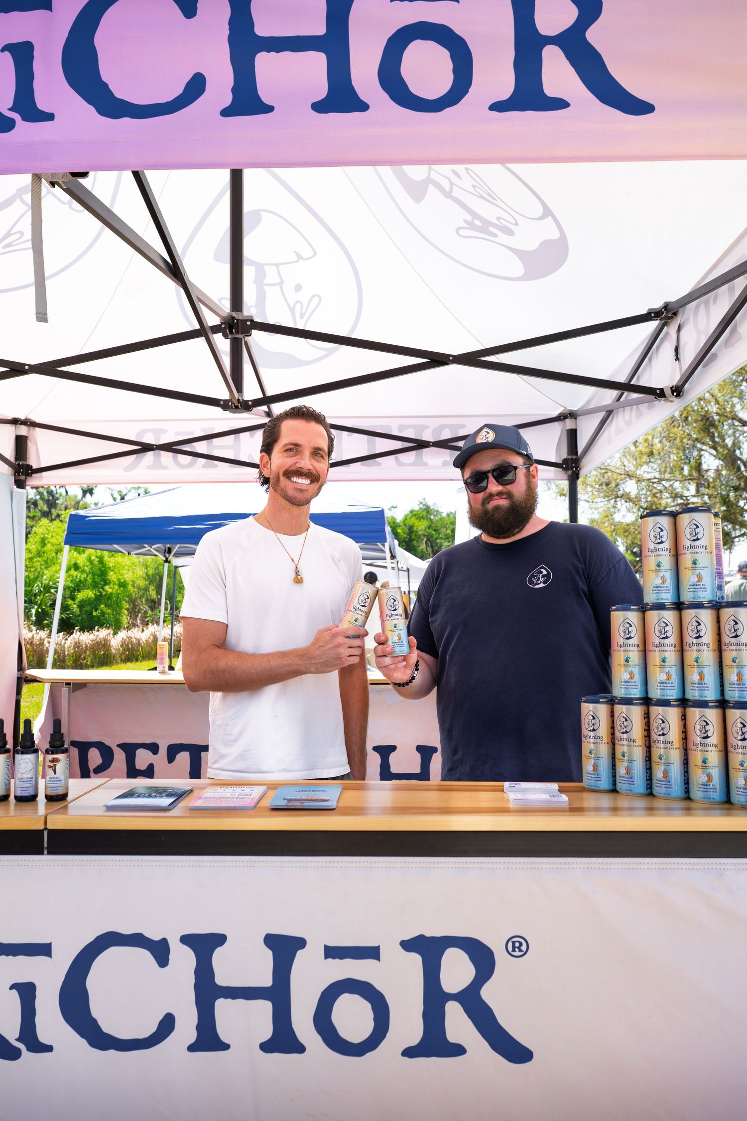 Two men smiling at an Anchor brand outdoor festival booth with canned beverages stacked behind them.