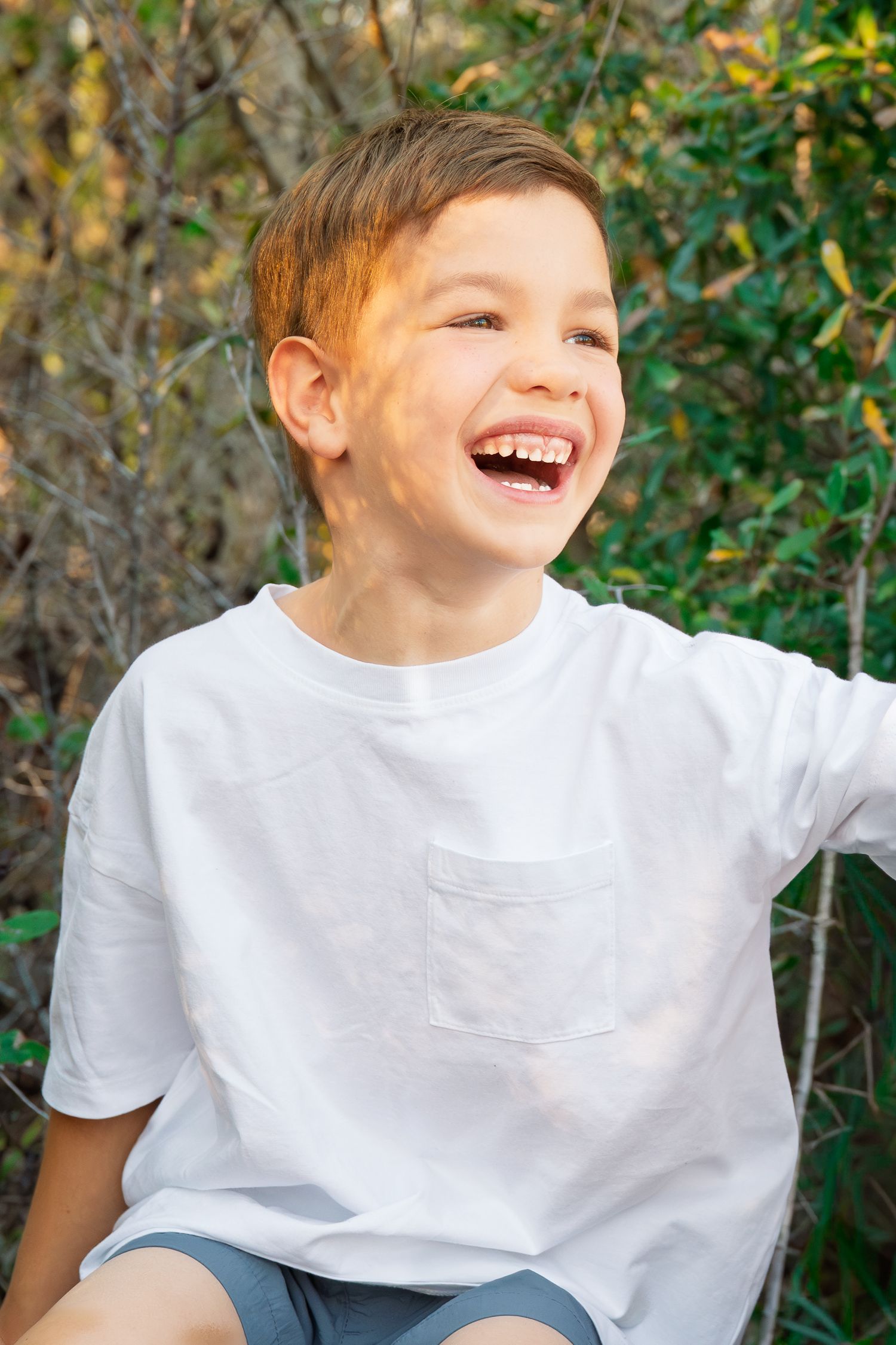 A joyful young boy in a white t-shirt laughs outdoors with green foliage in the background.