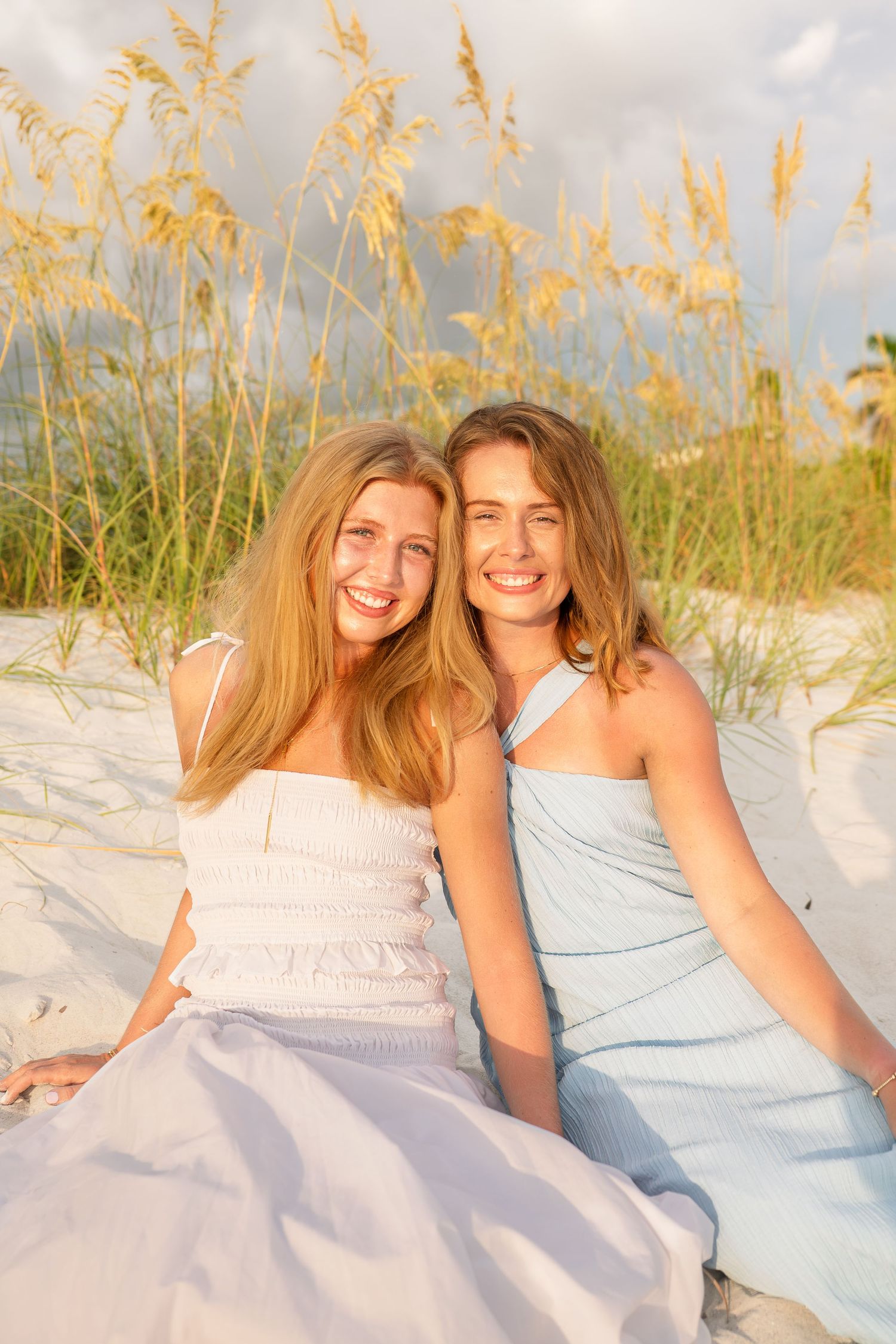 Two smiling young women in pastel dresses sitting on white sand dunes with sea oats at golden hour.