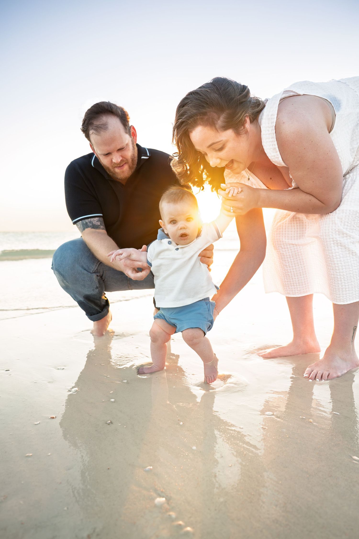 Young family helping baby take first steps on a sunny beach at golden hour.