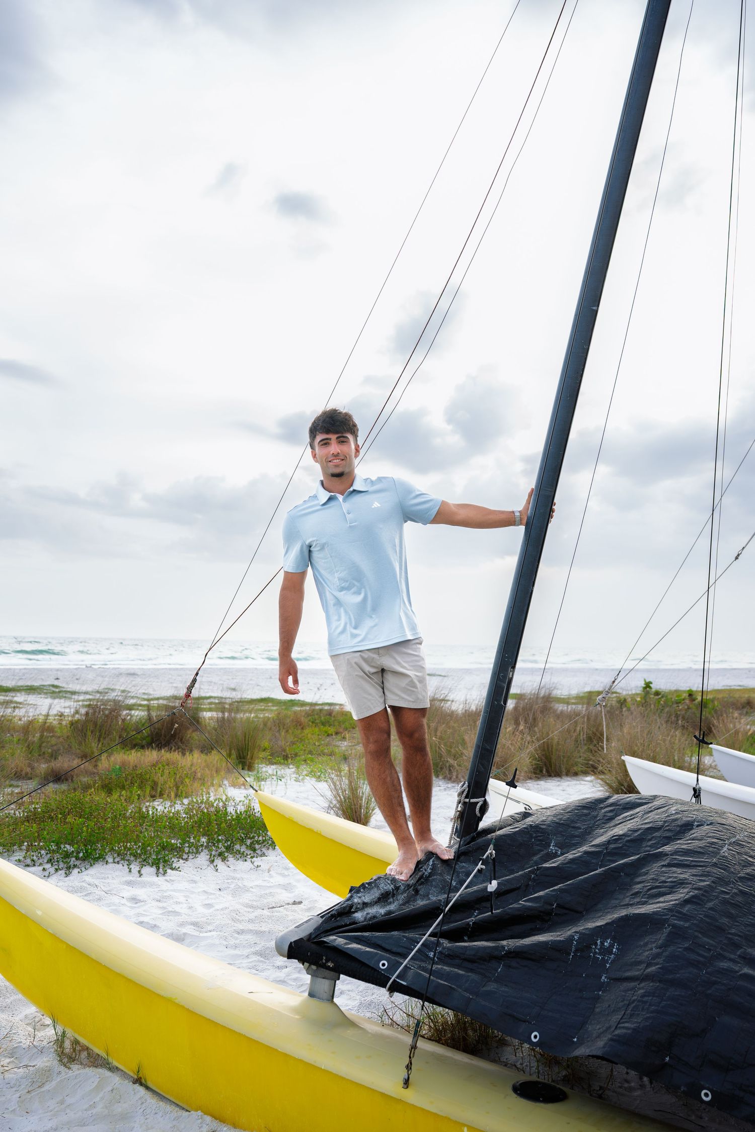 Young man standing on a yellow catamaran sailboat on a sandy beach with cloudy skies in the background.